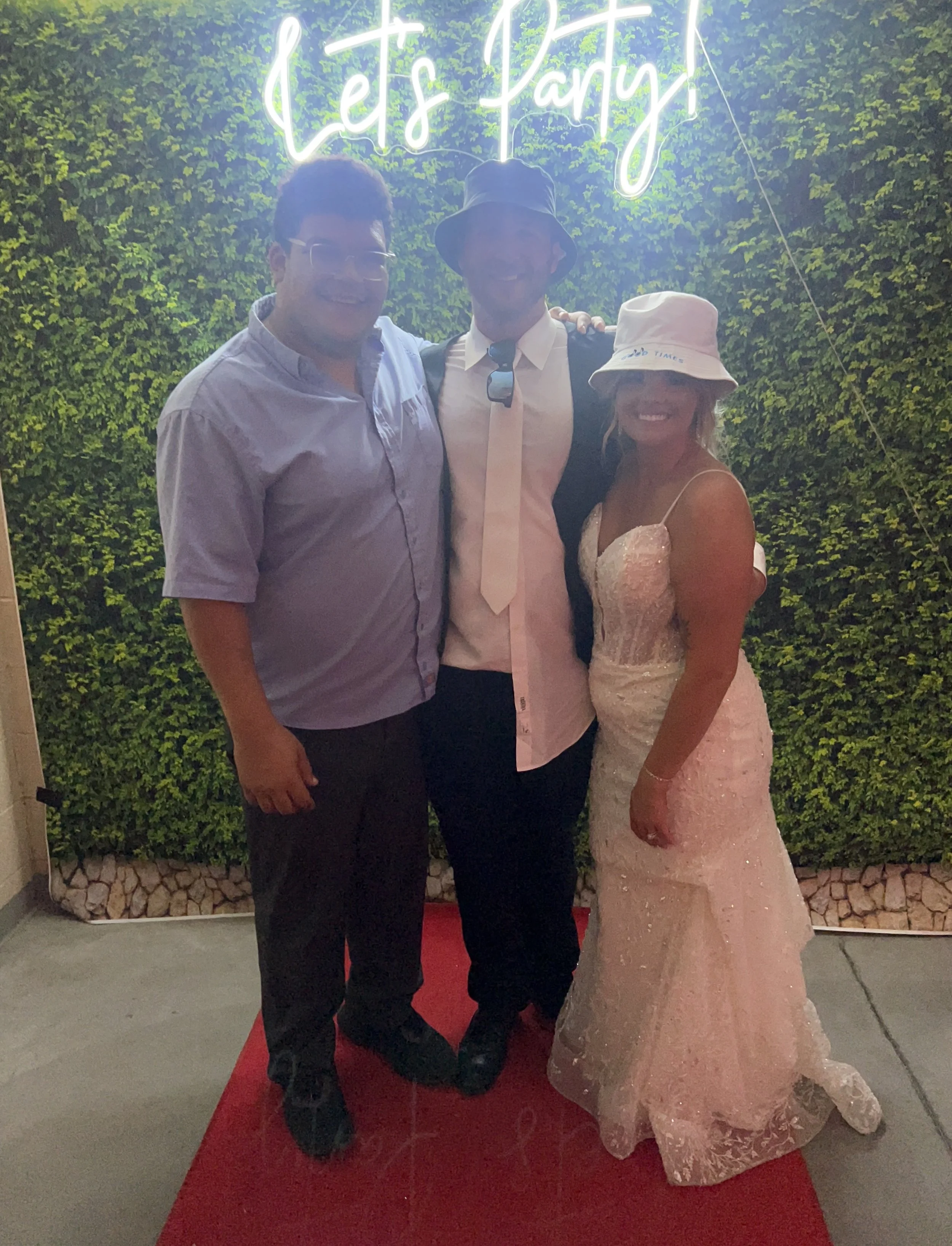 Three people standing on a red carpet in front of a green wall with a neon sign that says "Let's Party." The person on the left is wearing a light purple shirt and dark pants. The person in the middle is wearing a black hat, white shirt, black jacket