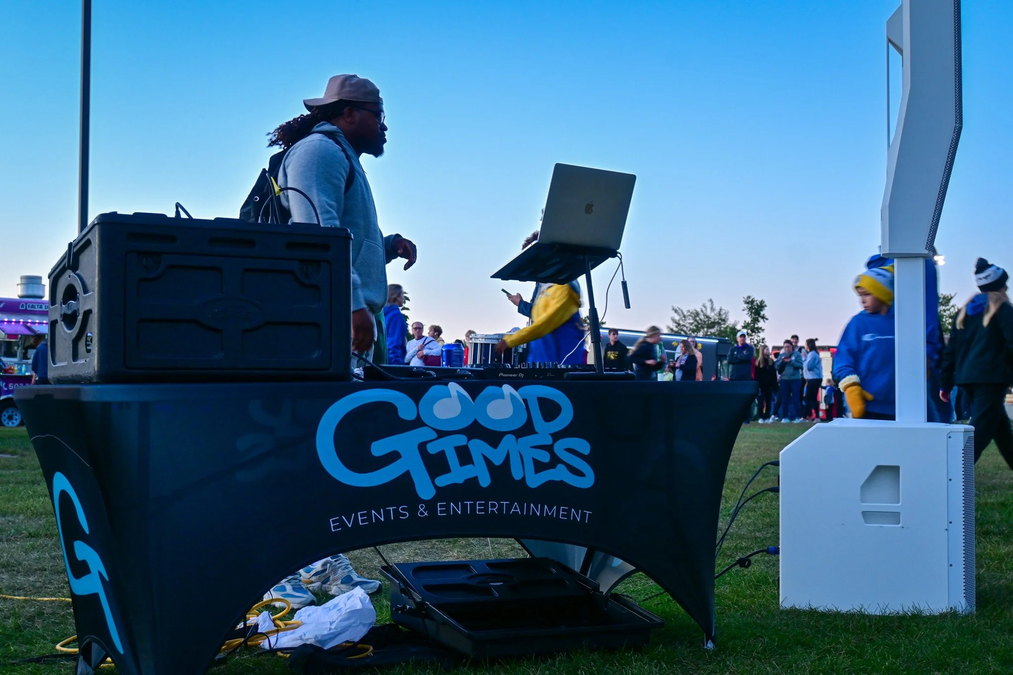 DJ Isaiah performing at an outdoor event, the Augie Twilight Run, with a crowd of people in the background, and a sign that reads 'Good Times Events & Entertainment' on the DJ booth.