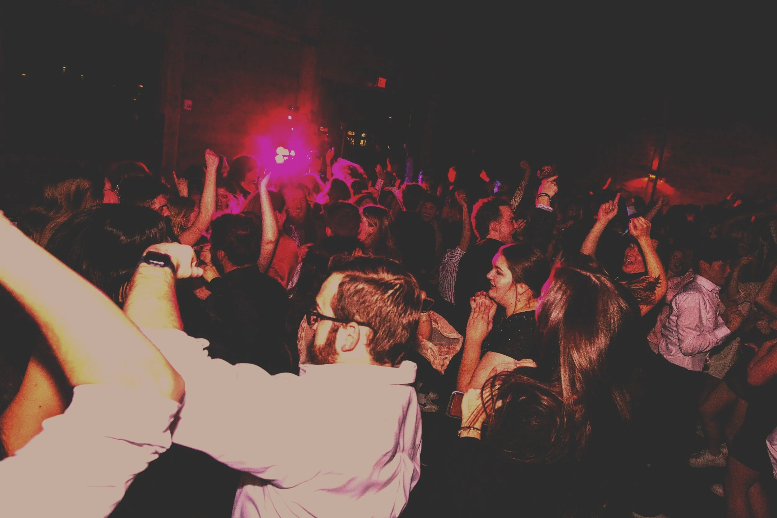 Crowd of people dancing and enjoying music at a nighttime party or concert, with pink and purple lighting.