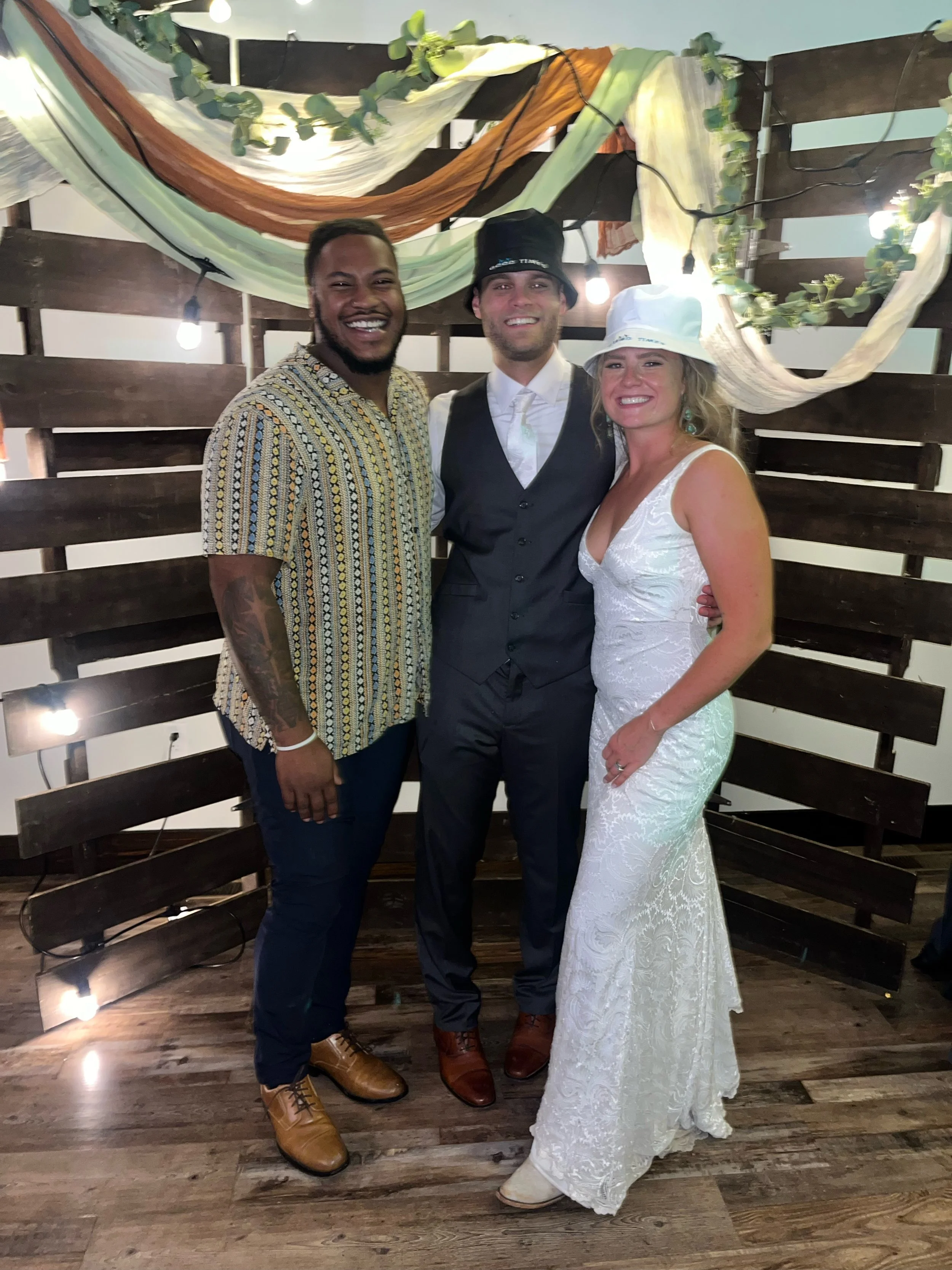Three people, two men and one woman, smiling and posing for a photo at a celebration or wedding, standing in front of a decorative wooden backdrop with fabric and string lights. The woman is wearing a white wedding dress and a white hat, while the me