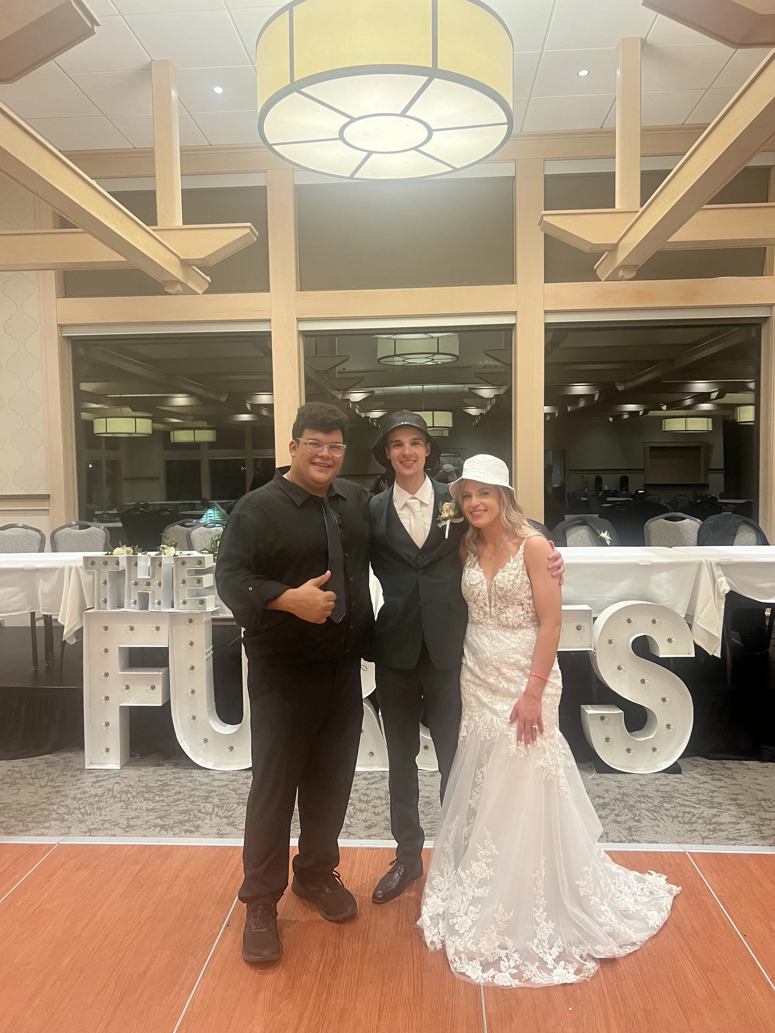 Three people standing together in formal attire at a wedding reception, with large decorative letters spelling 'HUES' in the background.
