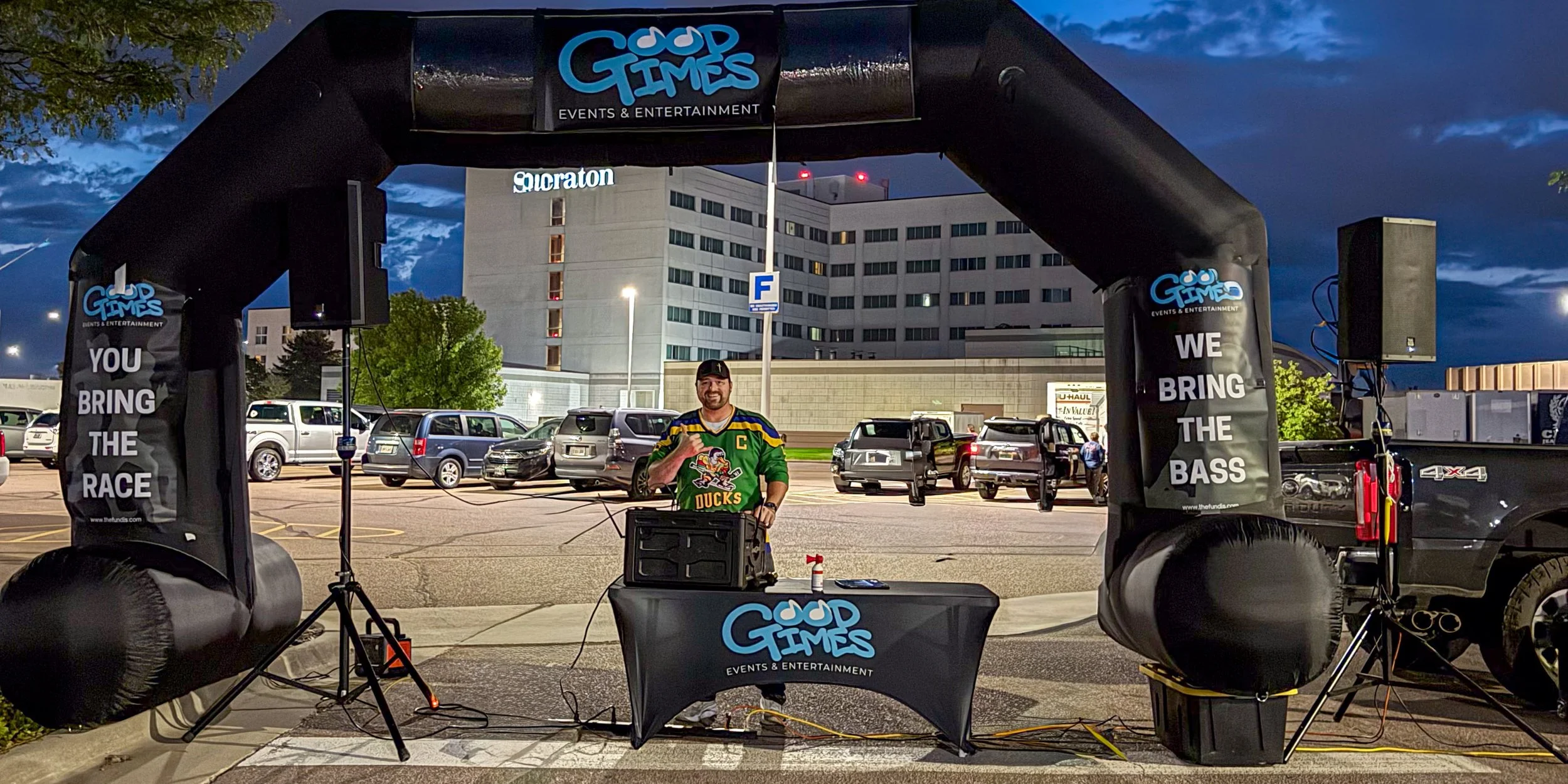 DJ Tony in a green hockey jersey standing behind a DJ booth at an outdoor event, the Sioux Falls Marathon, with cars and a building in the background during the early morning.