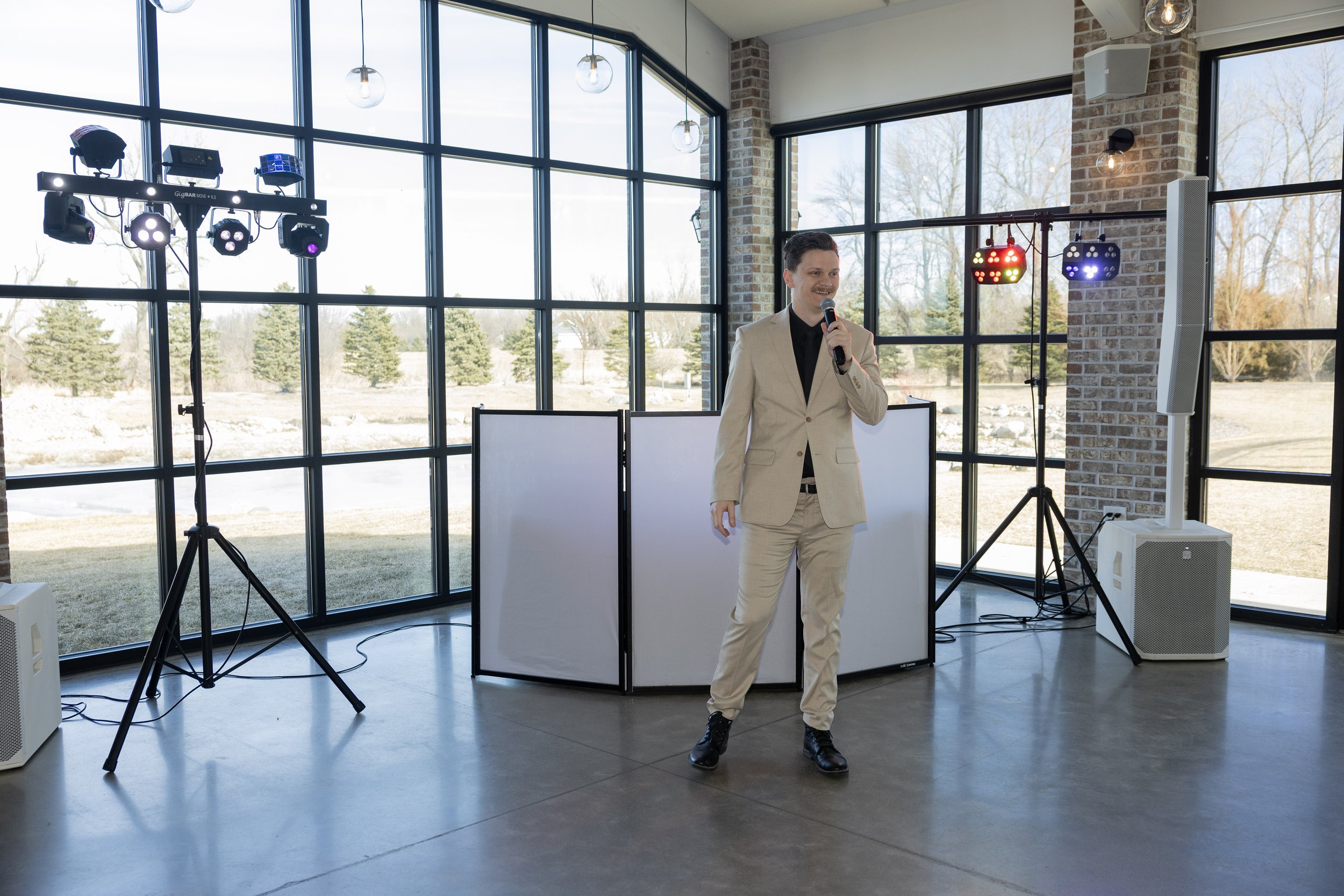 Man in beige suit holding microphone and speaking at indoor event with large windows, DJ equipment, and colorful lighting