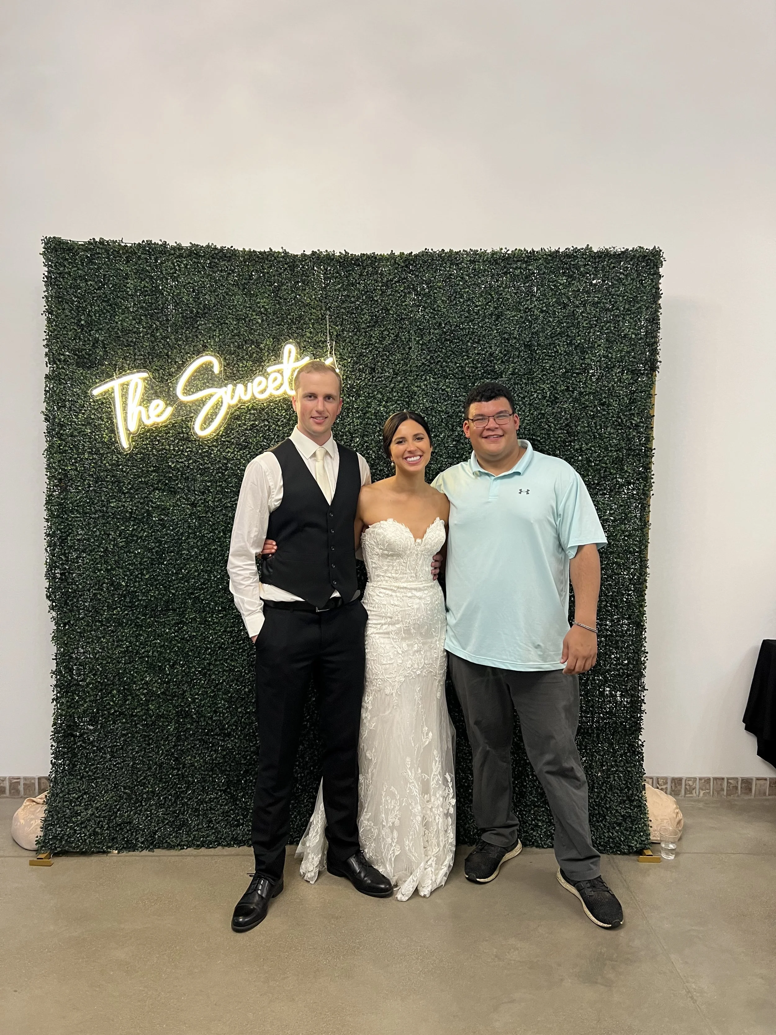 A bride and groom in wedding attire standing with a man in casual clothing in front of a green hedge wall with a neon sign that reads "The Sweet".
