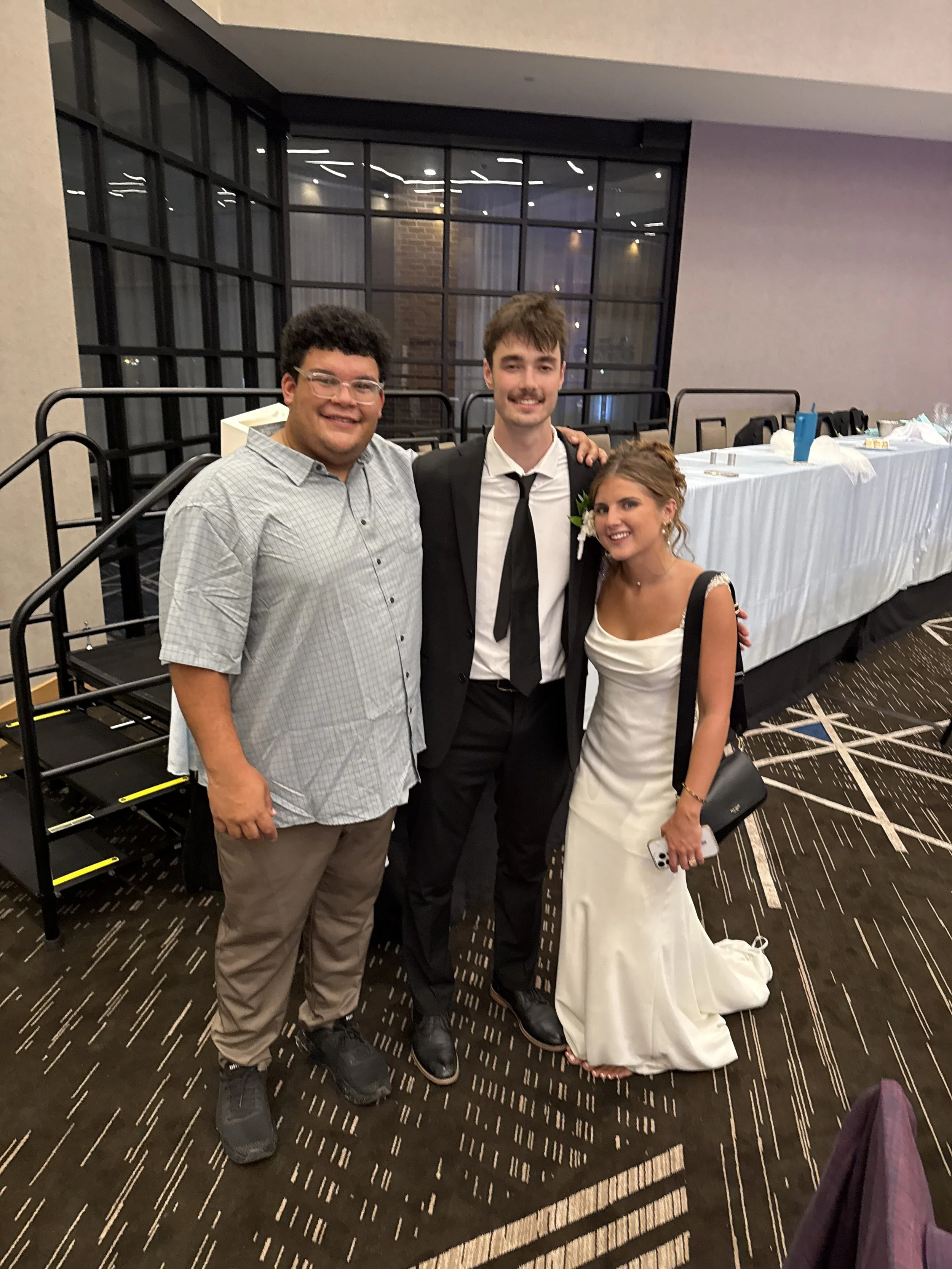 Three people smiling at a formal event, with two men and one woman dressed in a wedding gown. The background features a large window and a table covered with a white tablecloth.