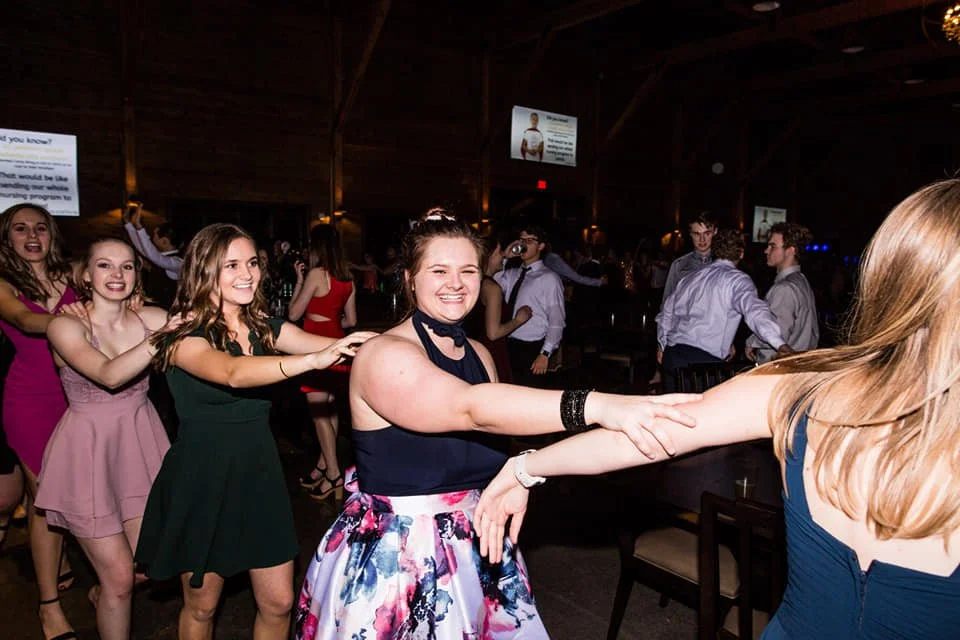 Group of young students dancing in a conga line at a school dance, smiling and holding each other's hands.