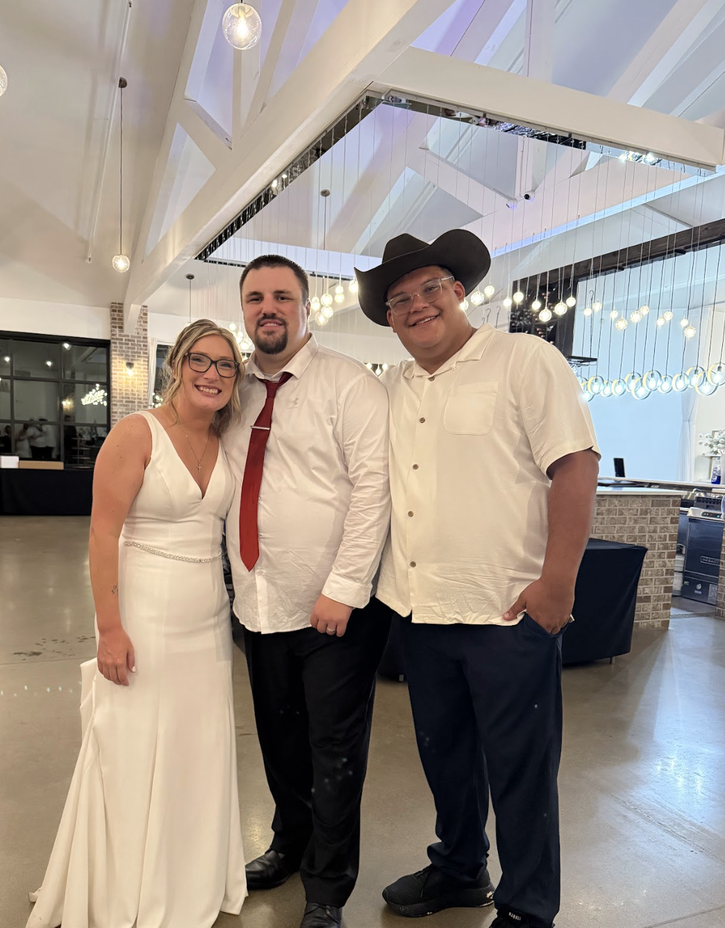 Three people standing together at a wedding reception, smiling. The woman on the left is wearing a white wedding dress and glasses. The man in the middle is wearing a white shirt, red tie, and dark pants. The man on the right is wearing a white shirt