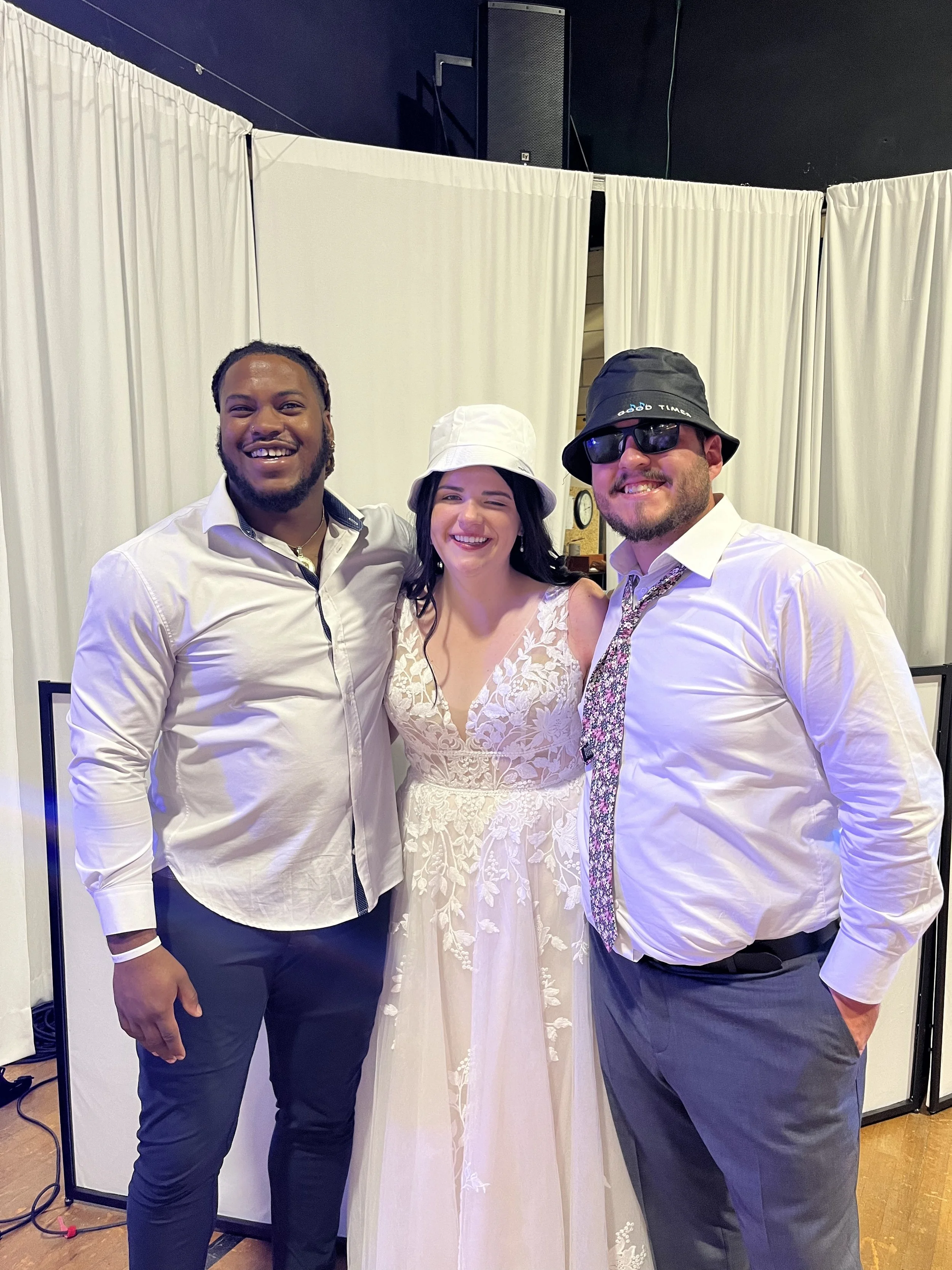 Three people in formal attire smiling at a celebration or wedding party, with white curtains behind them.