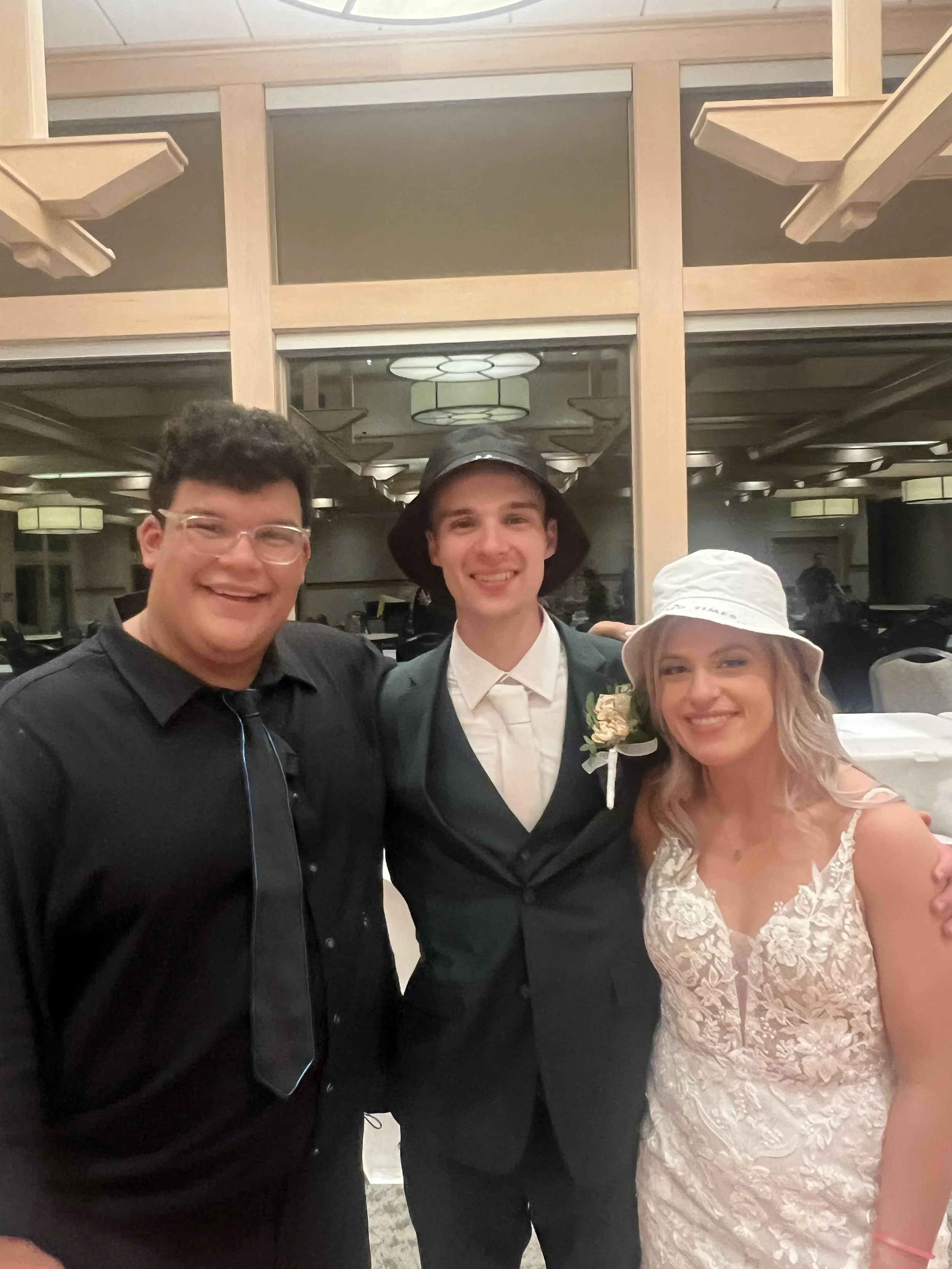 Three smiling people at a formal event, with two men and one woman, dressed in elegant attire. The man in the middle is wearing a dark suit with a white shirt and a hat, and the woman on the right is in a lace dress and a white hat. The man on the le