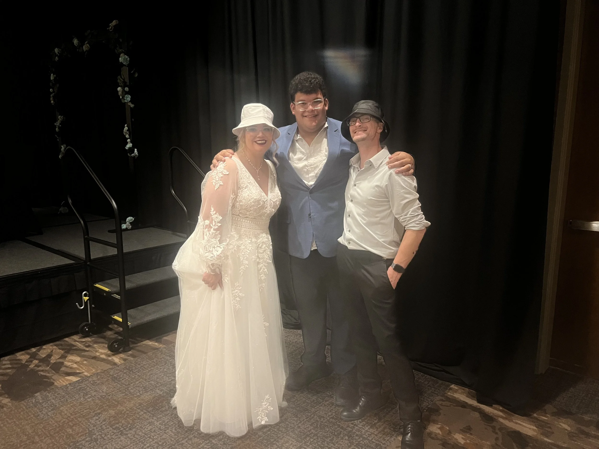 Three people posing together at an event, with two men and one woman, all dressed semi-formally or casually, standing on a patterned carpet in front of a black curtain backdrop.