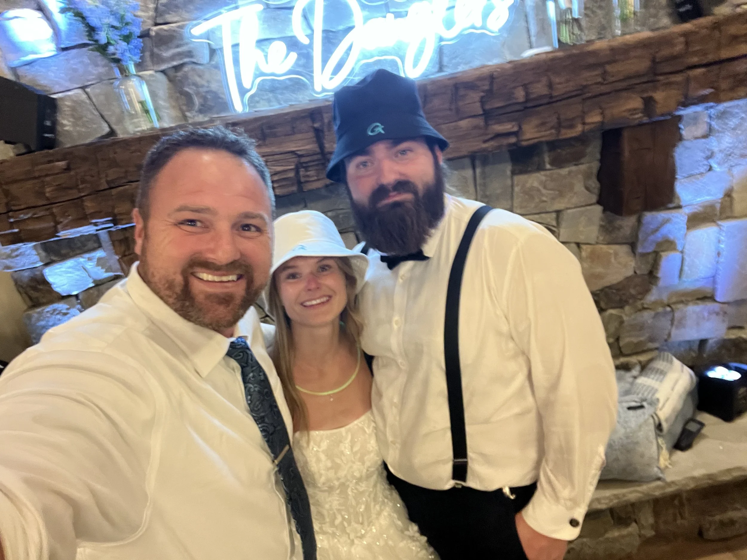 Three people taking a selfie at a party, with a stone wall and a neon sign in the background.