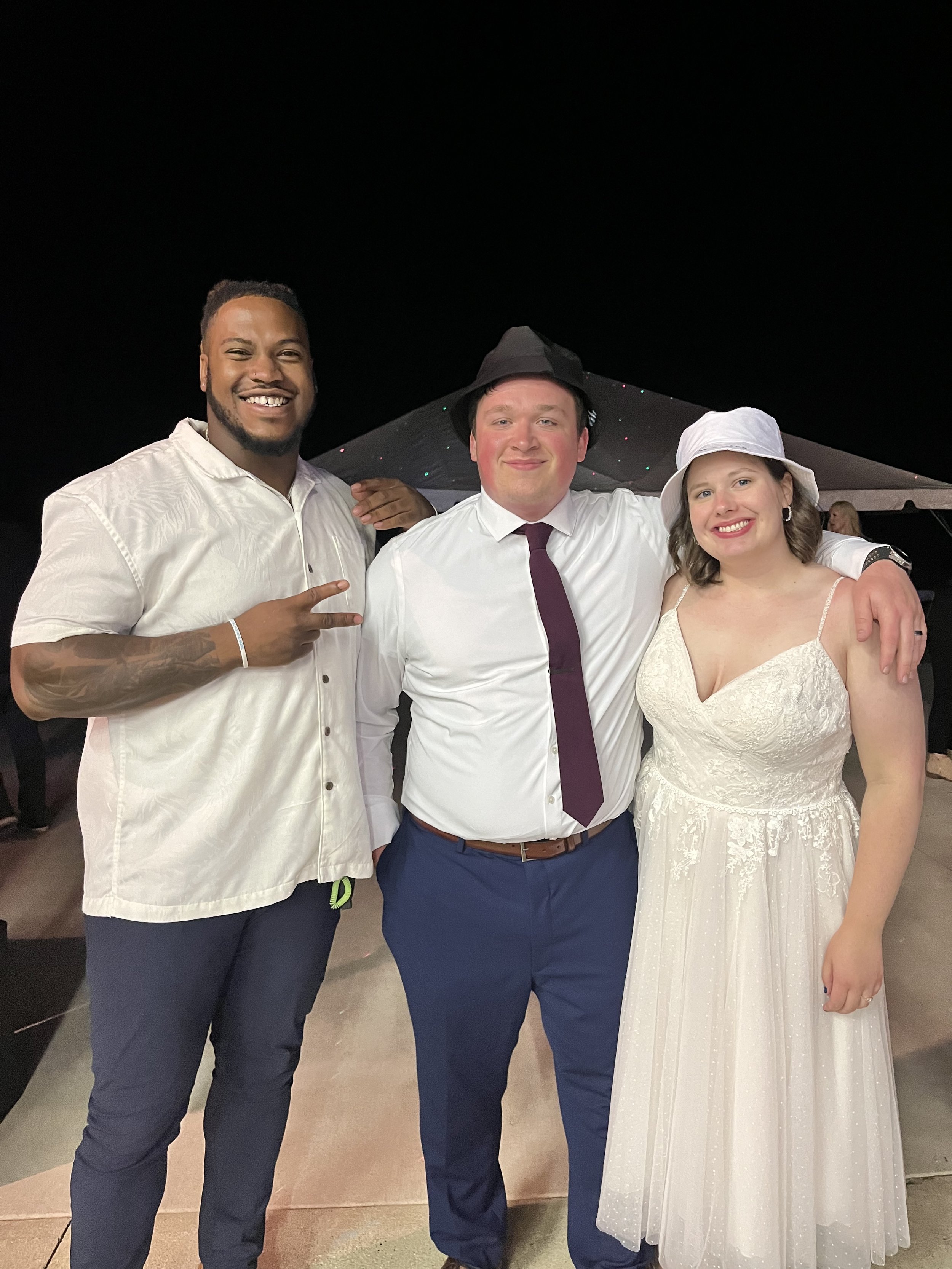 Three people at a nighttime celebration, with a man in the middle wearing a white shirt, purple tie, and black hat, flanked by a man on the left making a peace sign and a woman on the right in a white dress and white hat, all smiling.