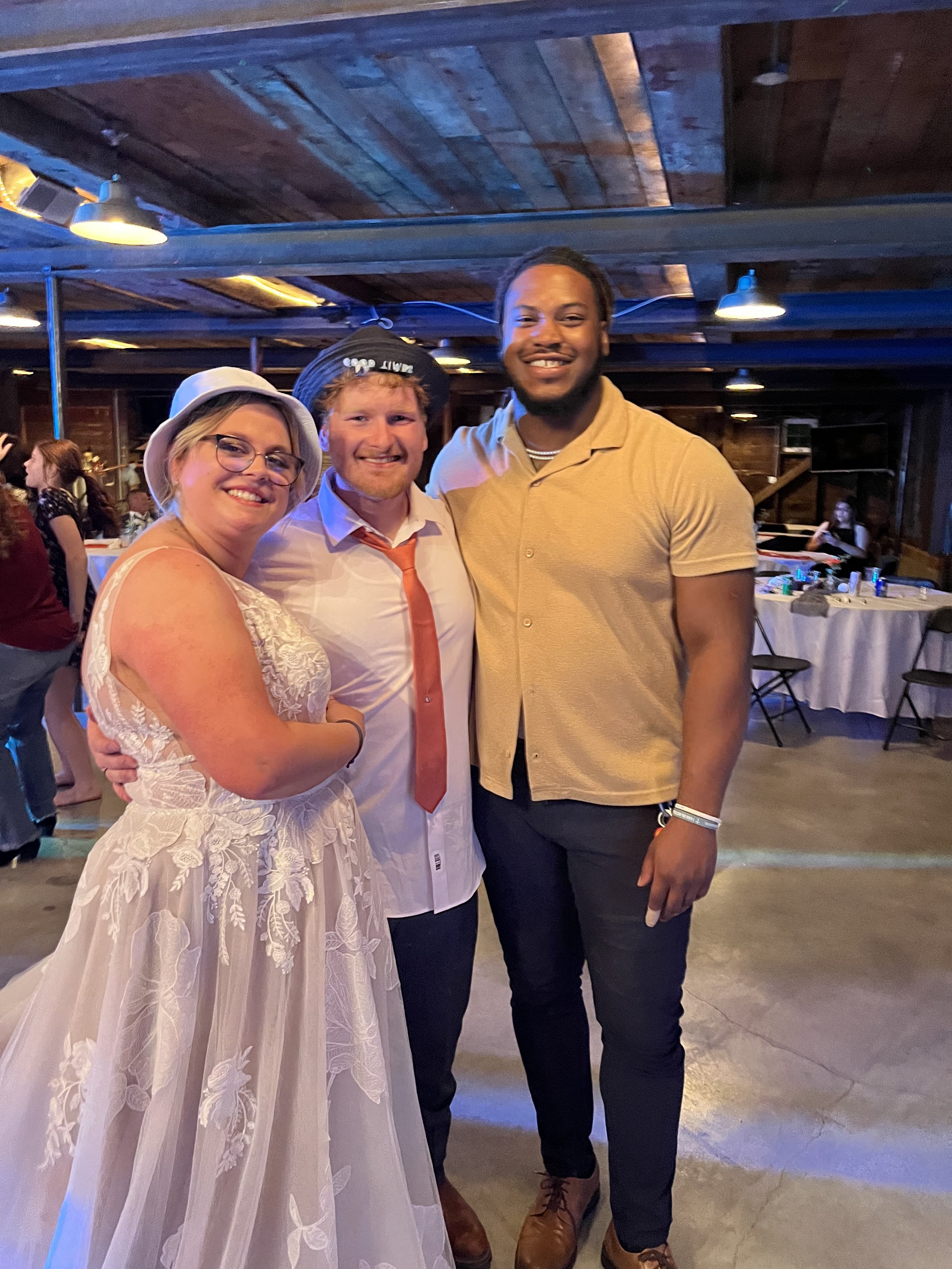 Three people standing together at a social event in a rustic wooden venue, smiling at the camera. The woman on the left is wearing a white lace dress and a white hat, the man in the middle is in a white shirt with a red tie and a beanie, and the man 