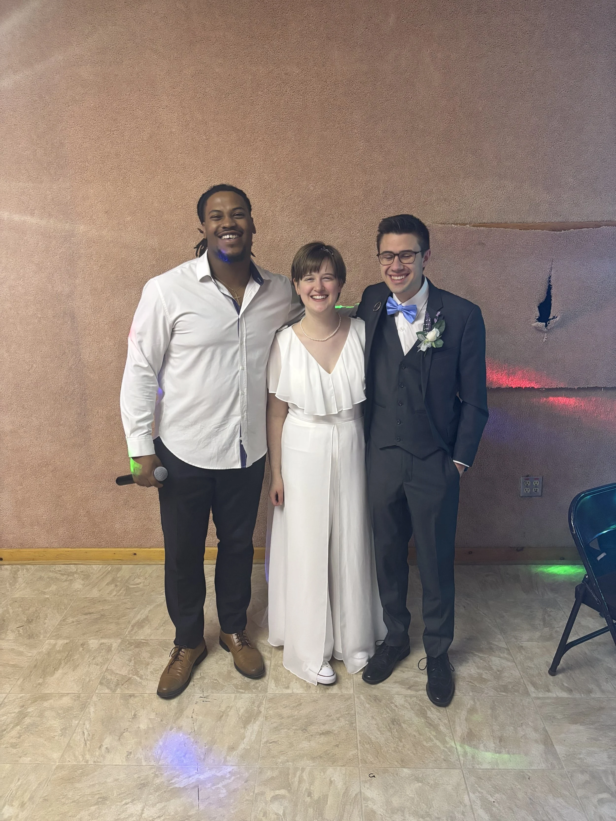 Three people are standing together at a celebration, smiling and dressed in formal attire. The person on the left is holding a microphone, the woman in the middle is wearing a white dress, and the man on the right is in a dark suit with a blue bow ti
