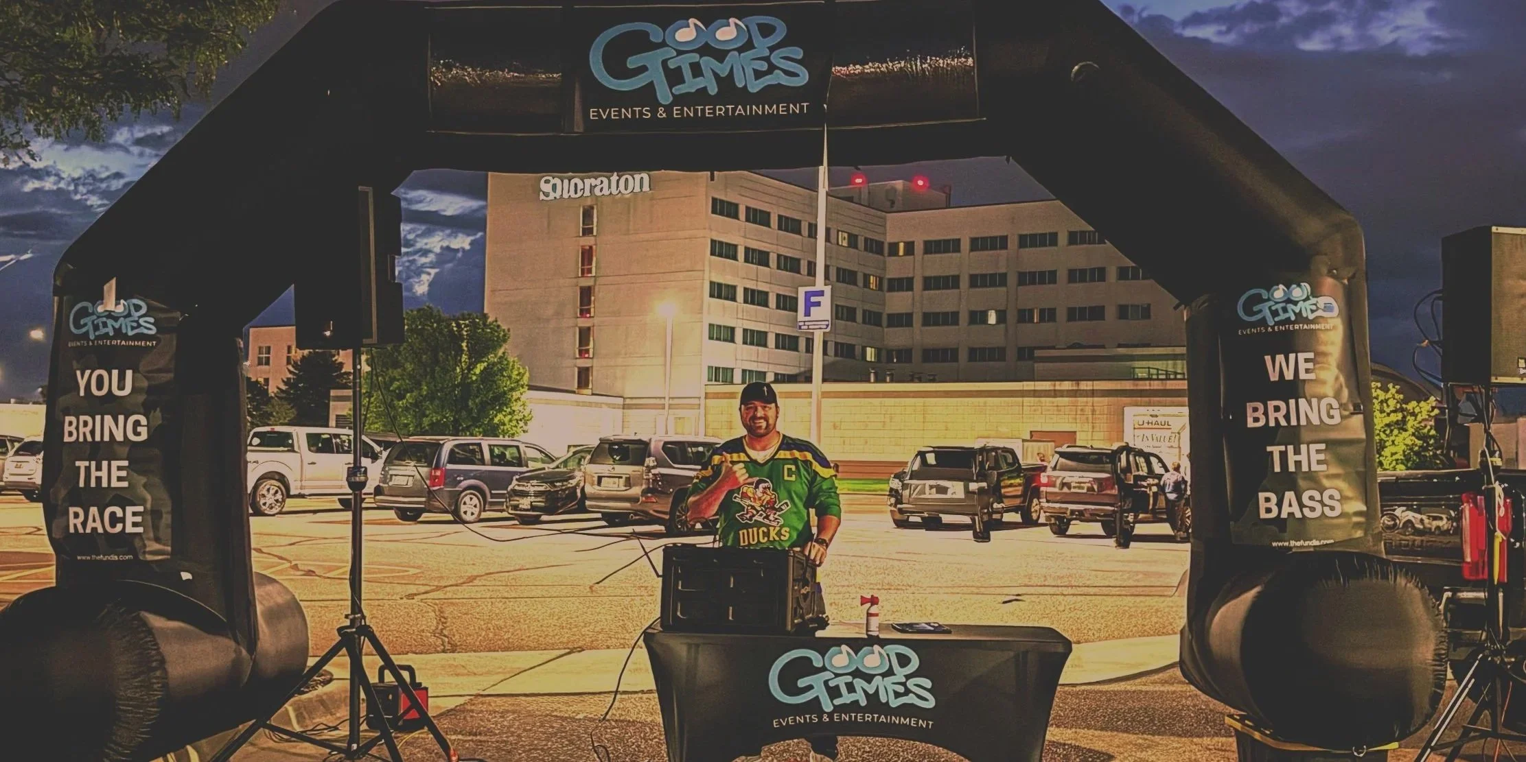 DJ Tony in a green hockey jersey standing behind a DJ booth at an outdoor event, the Sioux Falls Marathon, with cars and a building in the background during the early morning.