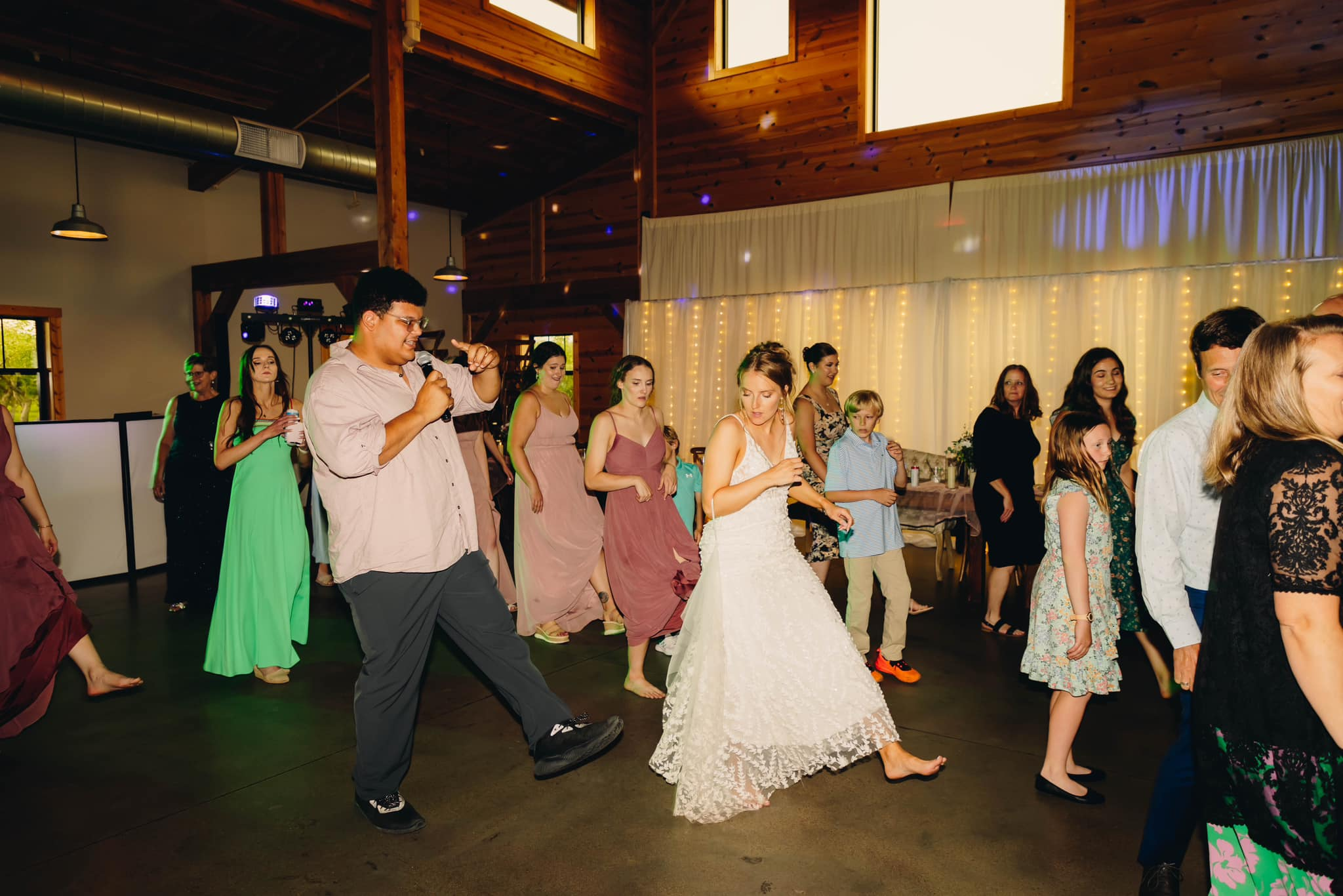 People dancing at a wedding reception in a rustic wooden barn with string lights and a white curtain backdrop.