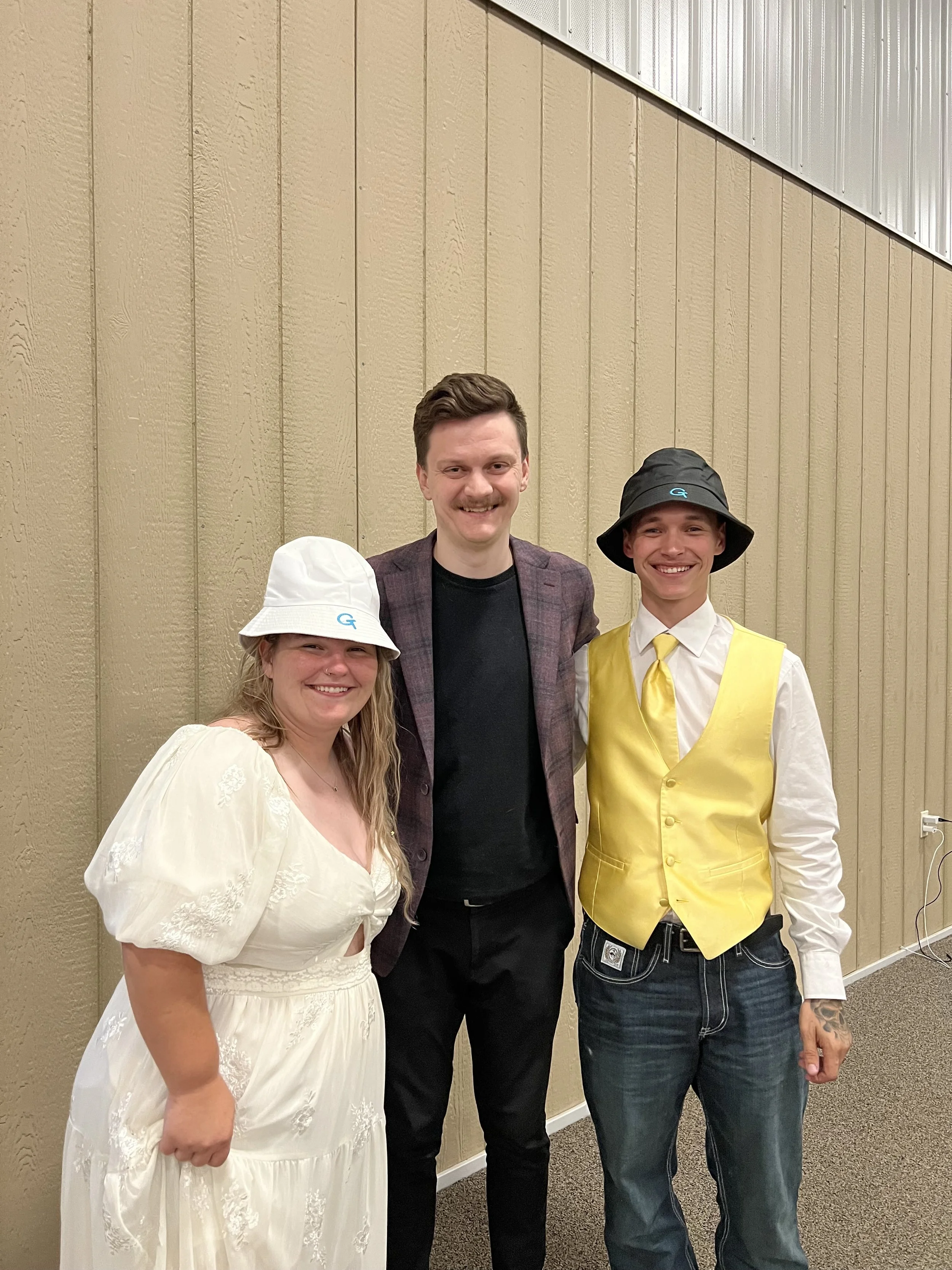 Three people standing together against a brown wall, smiling, dressed in casual and themed outfits. The woman on the left is wearing a white dress and white bucket hat, the man in the middle is wearing a blazer and black shirt, and the man on the rig