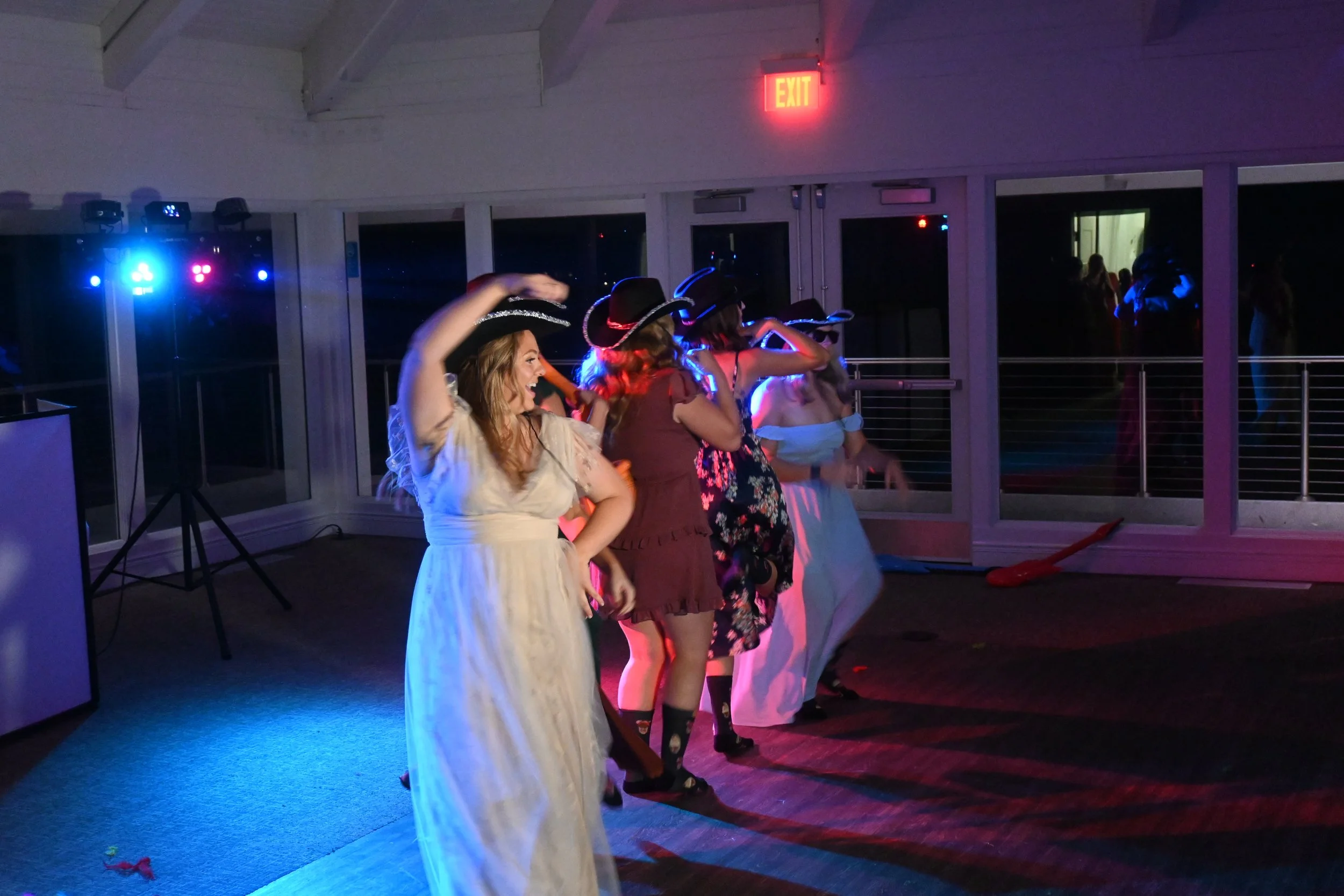 A group of women dressed in costumes, some wearing cowboy hats, dancing at a party or event in a decorated indoor venue with colorful lighting.