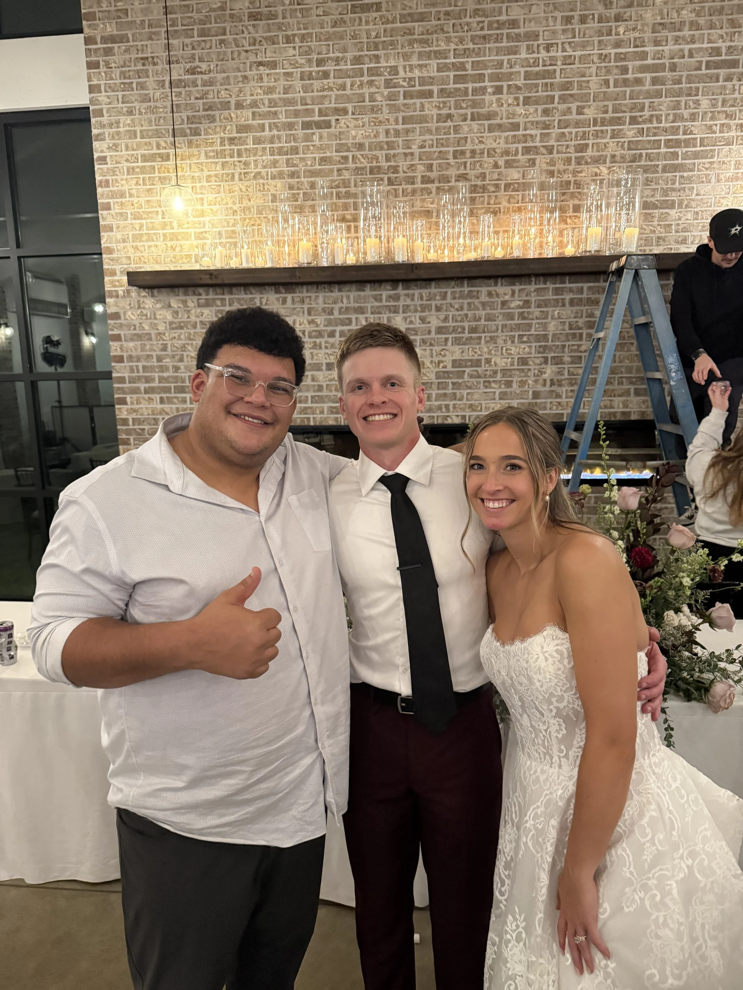Three people posing at a wedding reception. On the left is a man with glasses, wearing a light-colored shirt, giving a thumbs-up. In the middle is a man in a white shirt and black tie, smiling. On the right is a woman in a white lace wedding dress, s