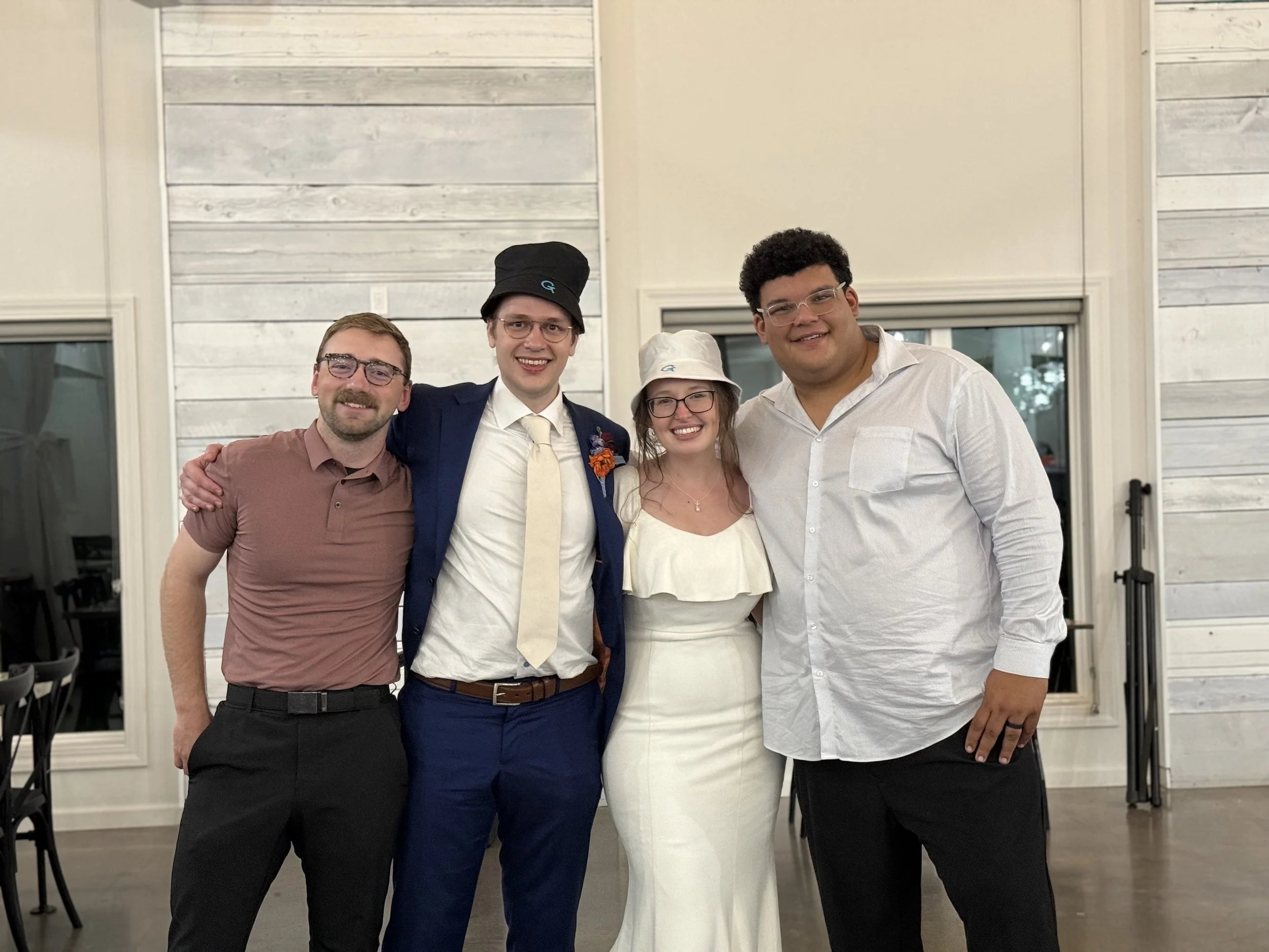 Four people smiling and standing close together indoors in front of a white wooden wall, celebrating an event, with large windows and black chairs visible in the background.