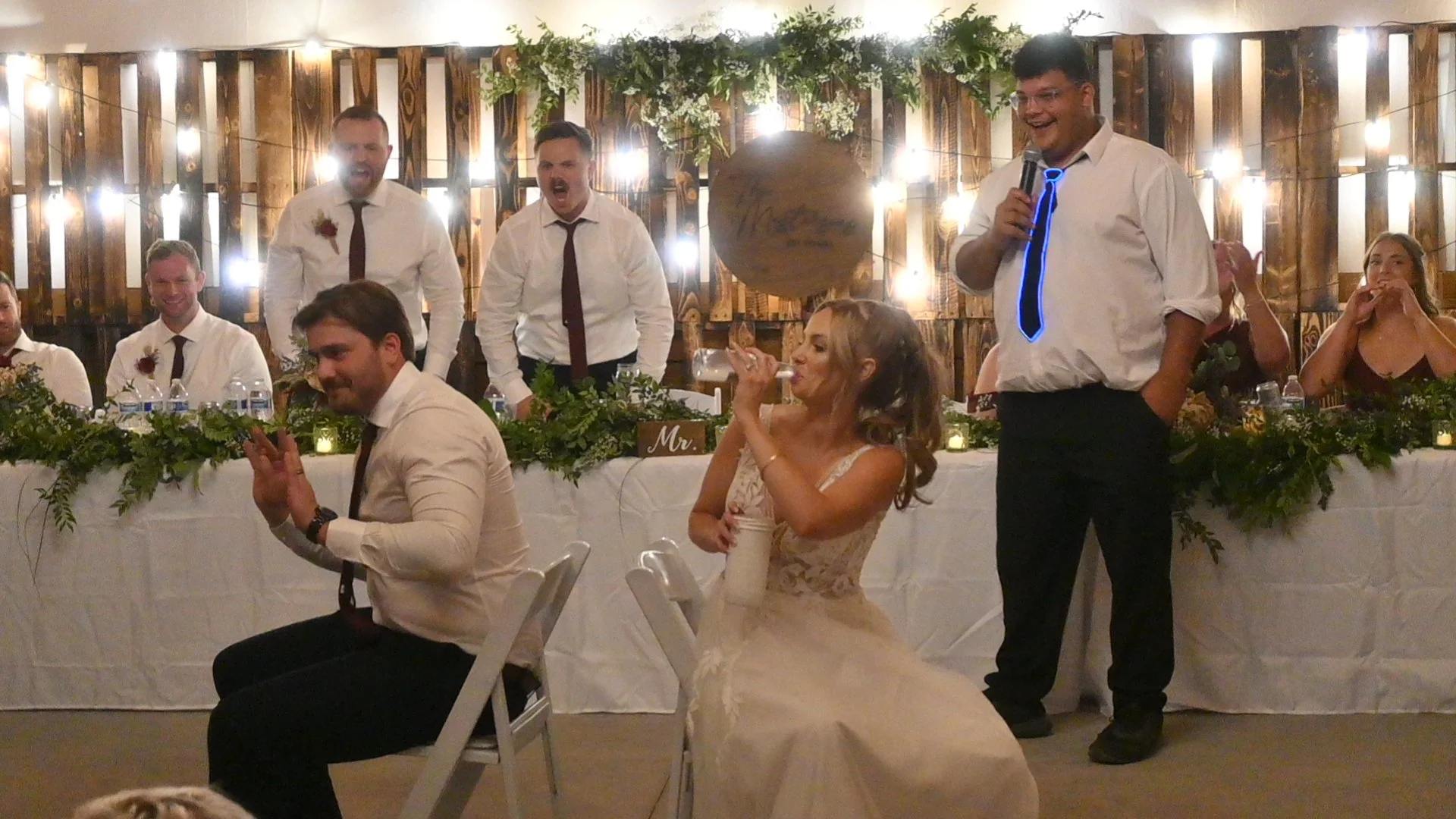 A wedding reception with a bridal party and guests. The bride, in a wedding dress, is drinking from a glass, while a man standing to her right is giving a speech or toast with a microphone. Several men in white shirts and ties are seated and standing
