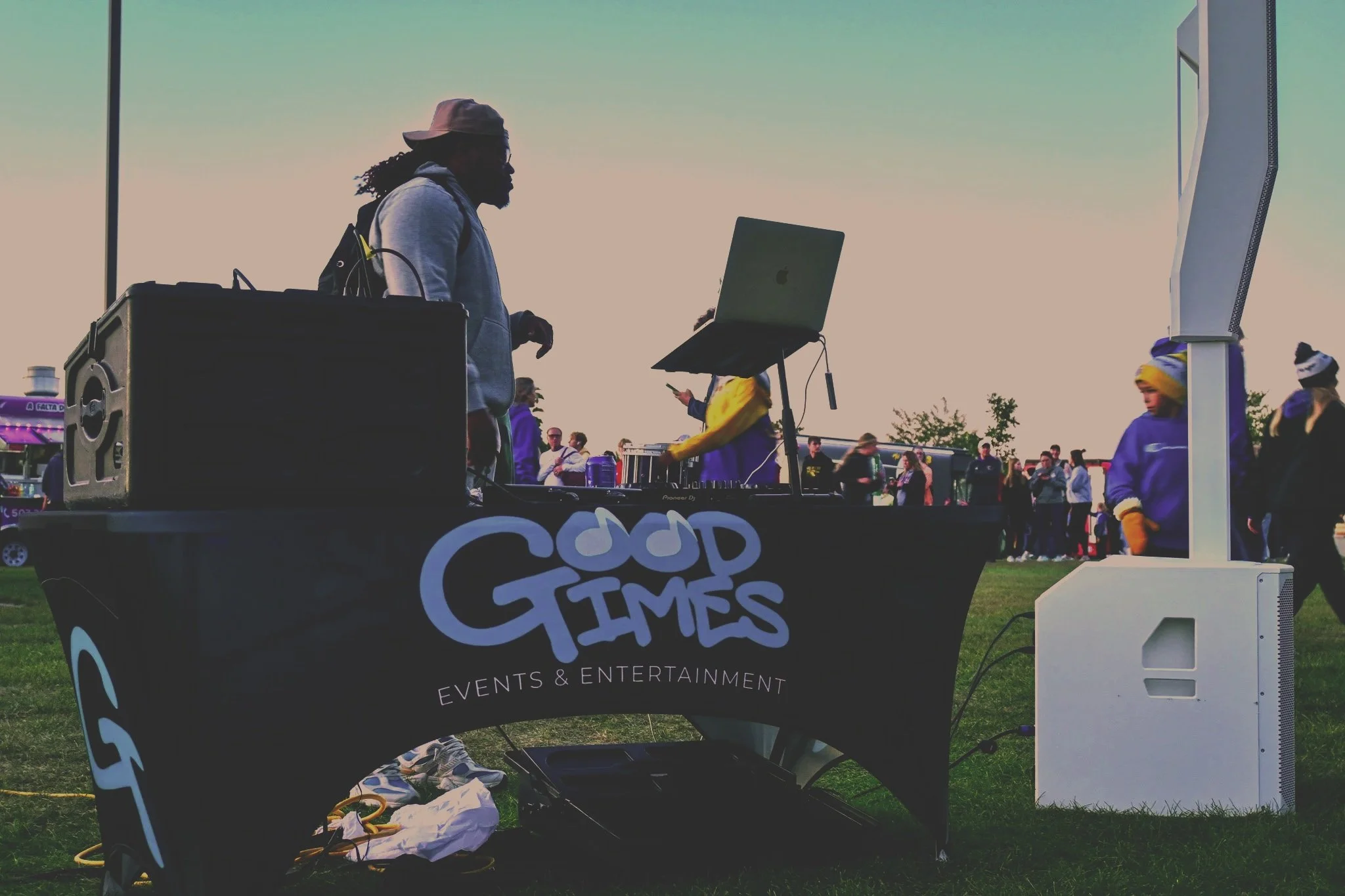 DJ Isaiah performing at an outdoor event, the Augie Twilight Run, with a crowd of people in the background, and a sign that reads 'Good Times Events & Entertainment' on the DJ booth.