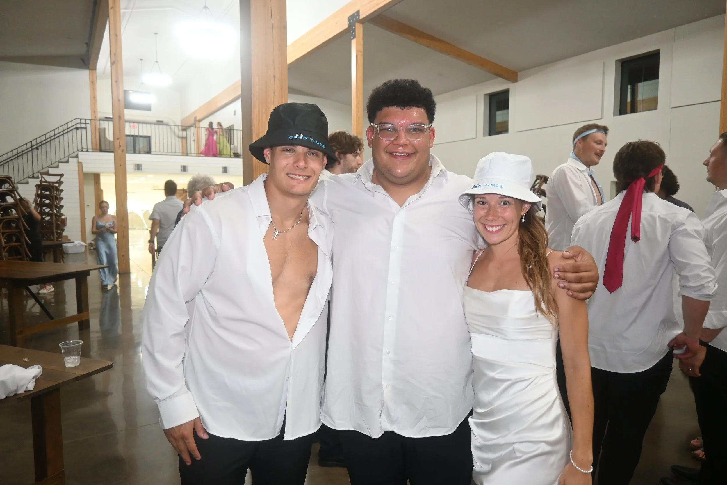 Three people standing together at an indoor event, smiling for the camera. The person on the left is a young man wearing a white shirt, open at the chest, with a black hat and a silver cross necklace. The person in the middle is a man in a white shir