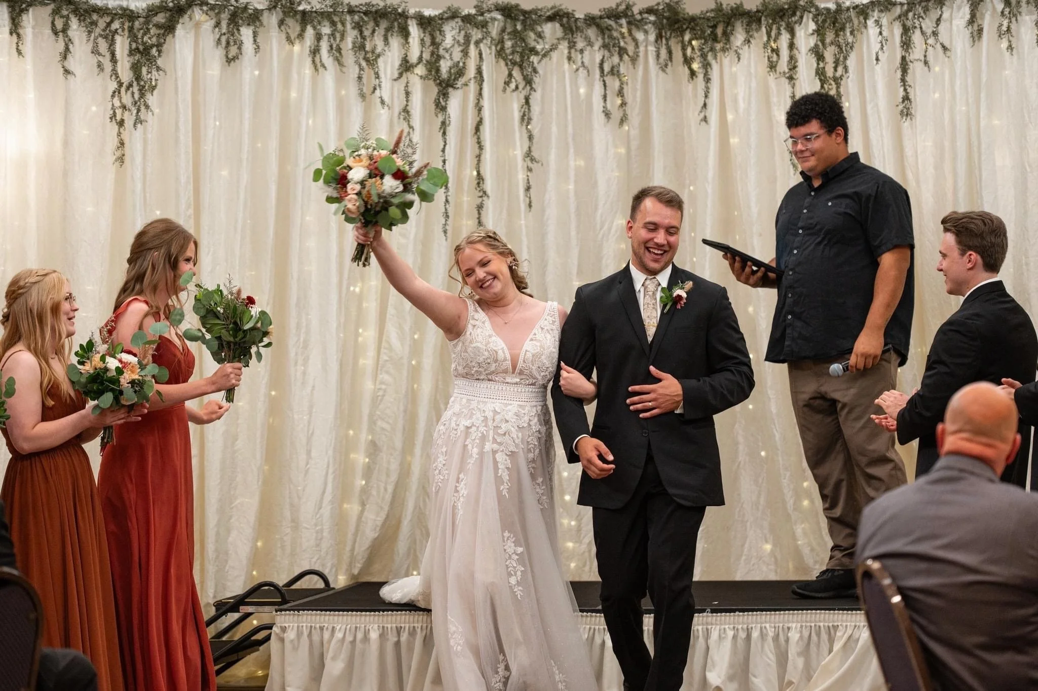 A bride and groom celebrate as they walk down a stage during their wedding reception, surrounded by bridesmaids and groomsmen, with a decorated curtain backdrop.