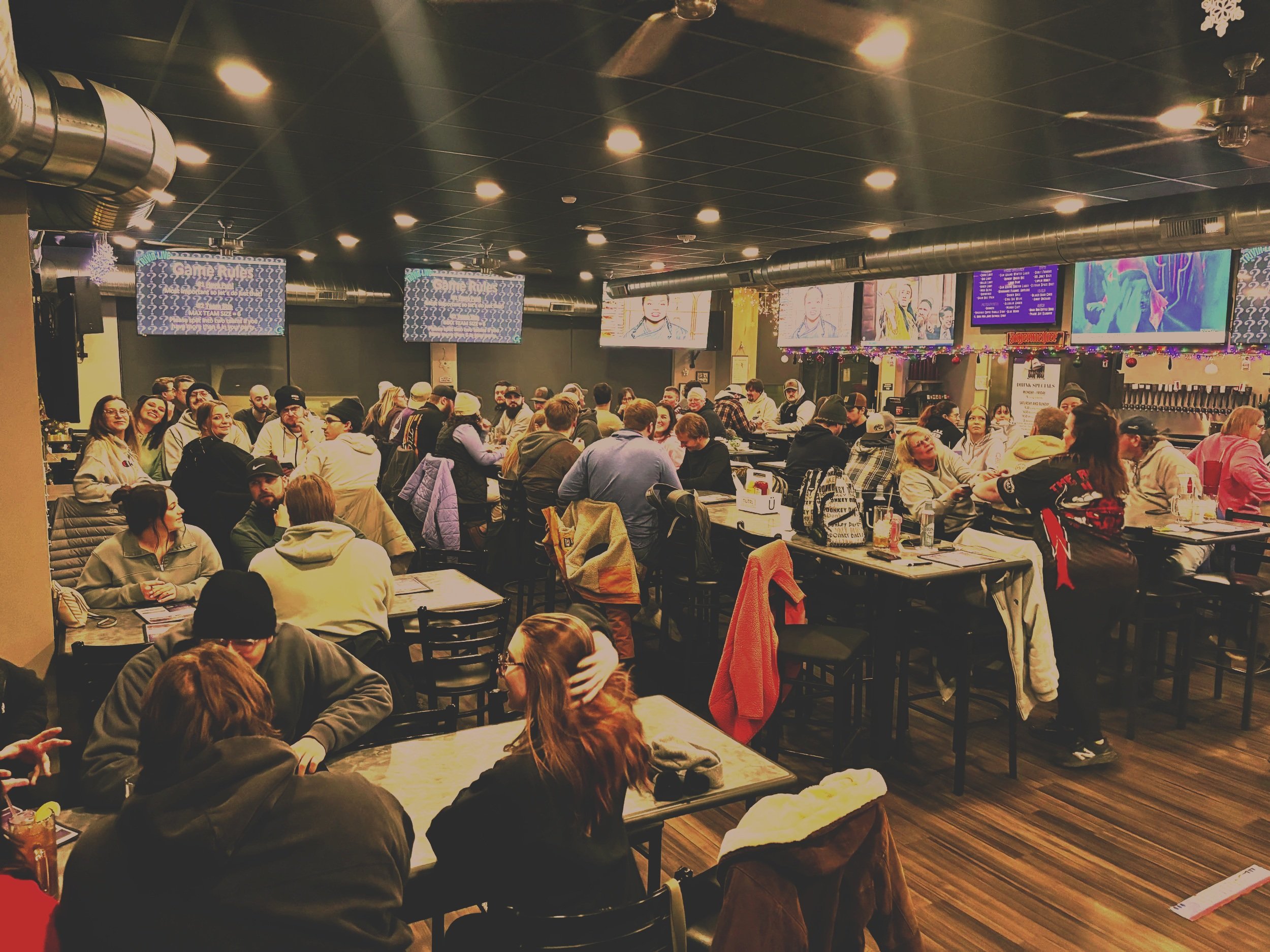 Crowded bar or restaurant with groups of people sitting at tables, watching TV screens on the wall, and playing Good Times Live Trivia.