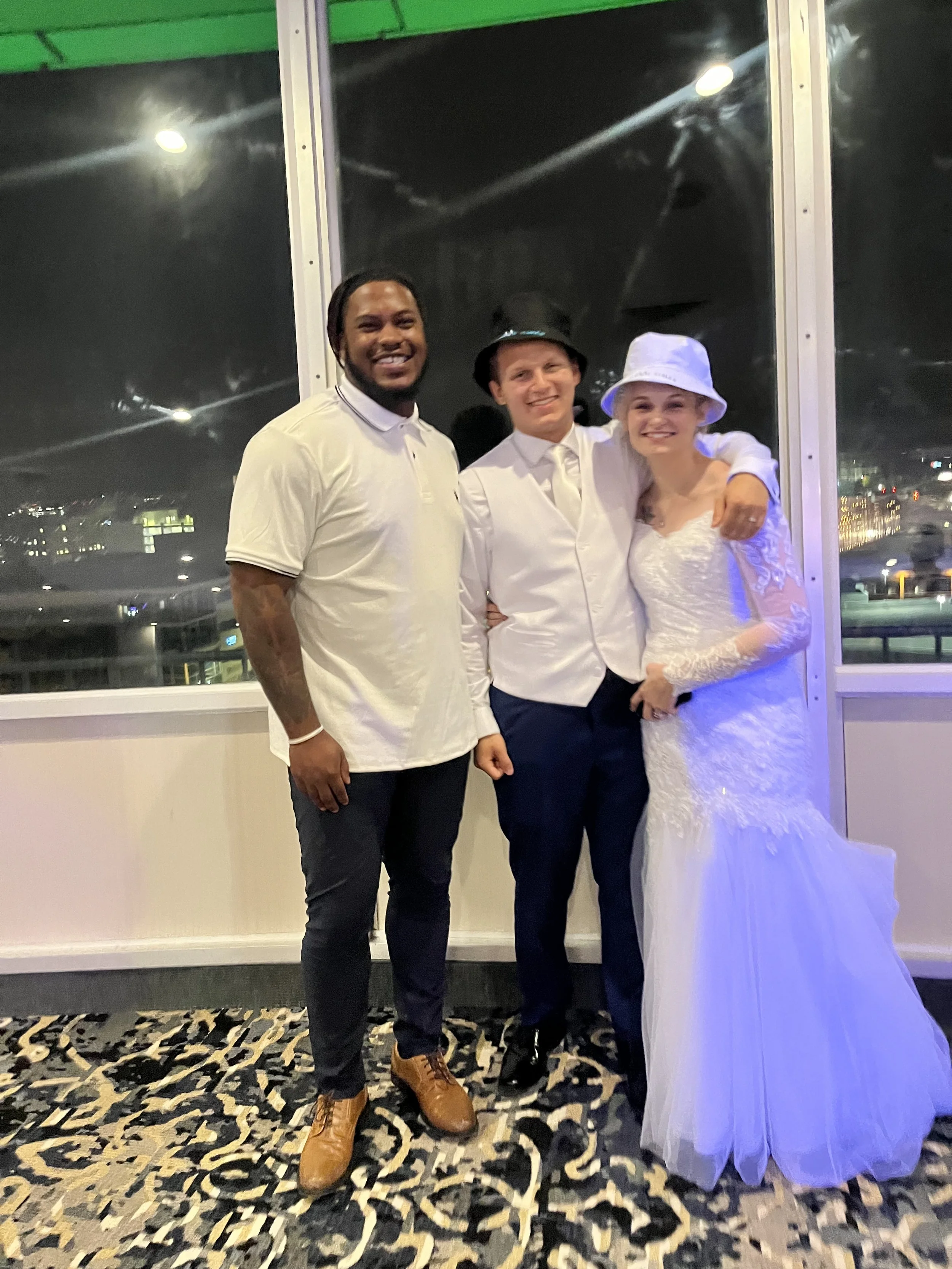 Three people, two men and one woman, standing together indoors at night, dressed in casual and wedding attire, smiling for the camera.