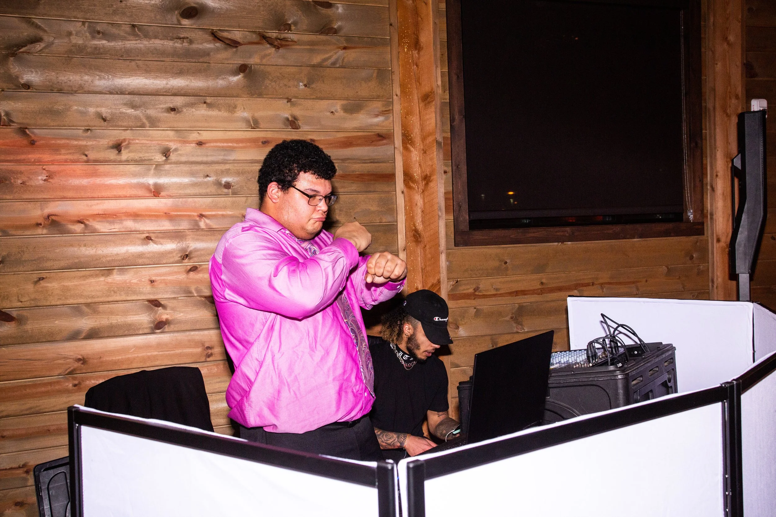 A man with curly hair, glasses, and a pink jacket stands with arms crossed in front of a DJ at a booth with equipment, inside a room with wooden walls and a large black television.
