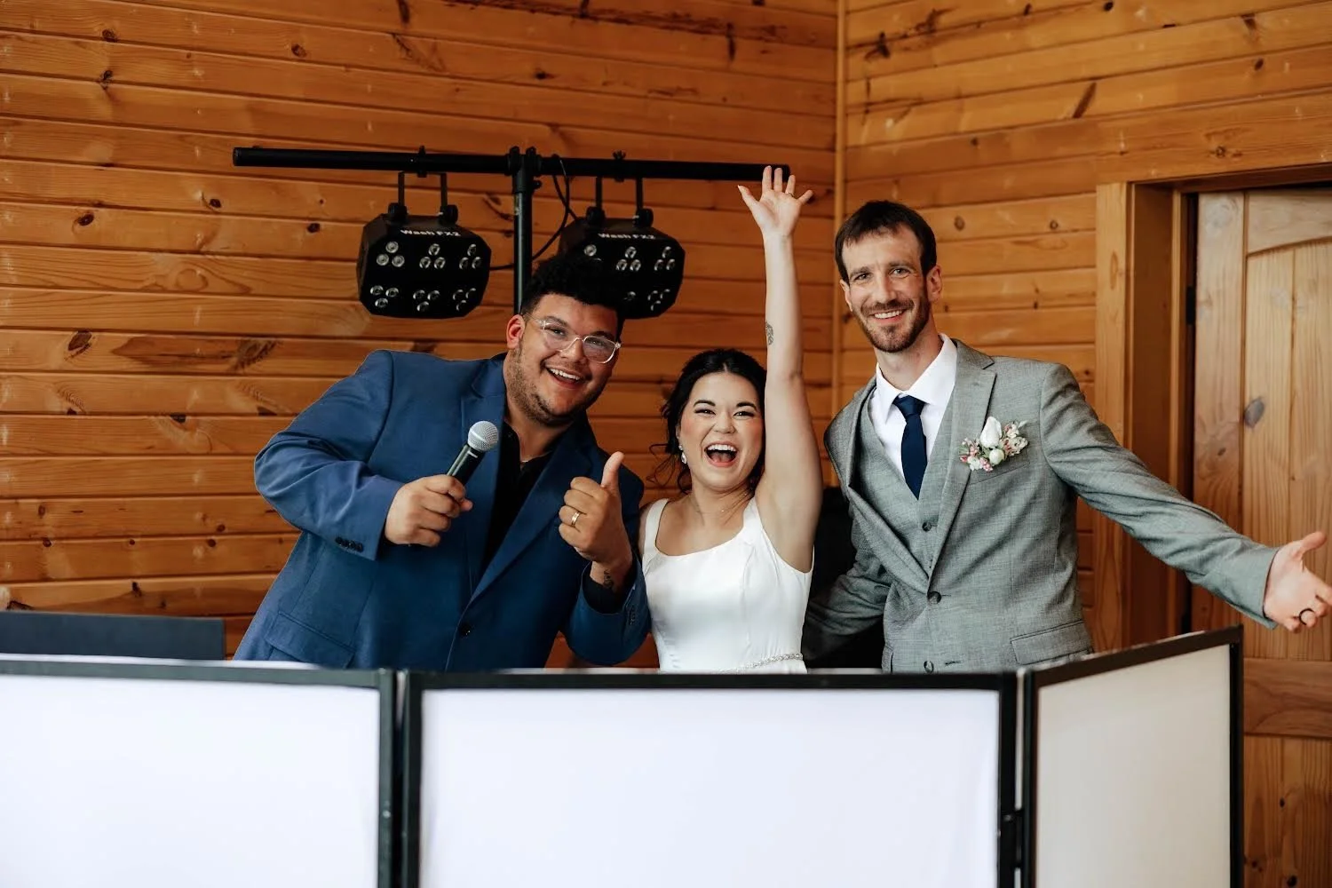 DJ Logan and a couple celebrating at a wedding, with a woman in a white dress in the center, with by two men in suits, one holding a microphone and the other with his arms open, in front of a wood-paneled wall.