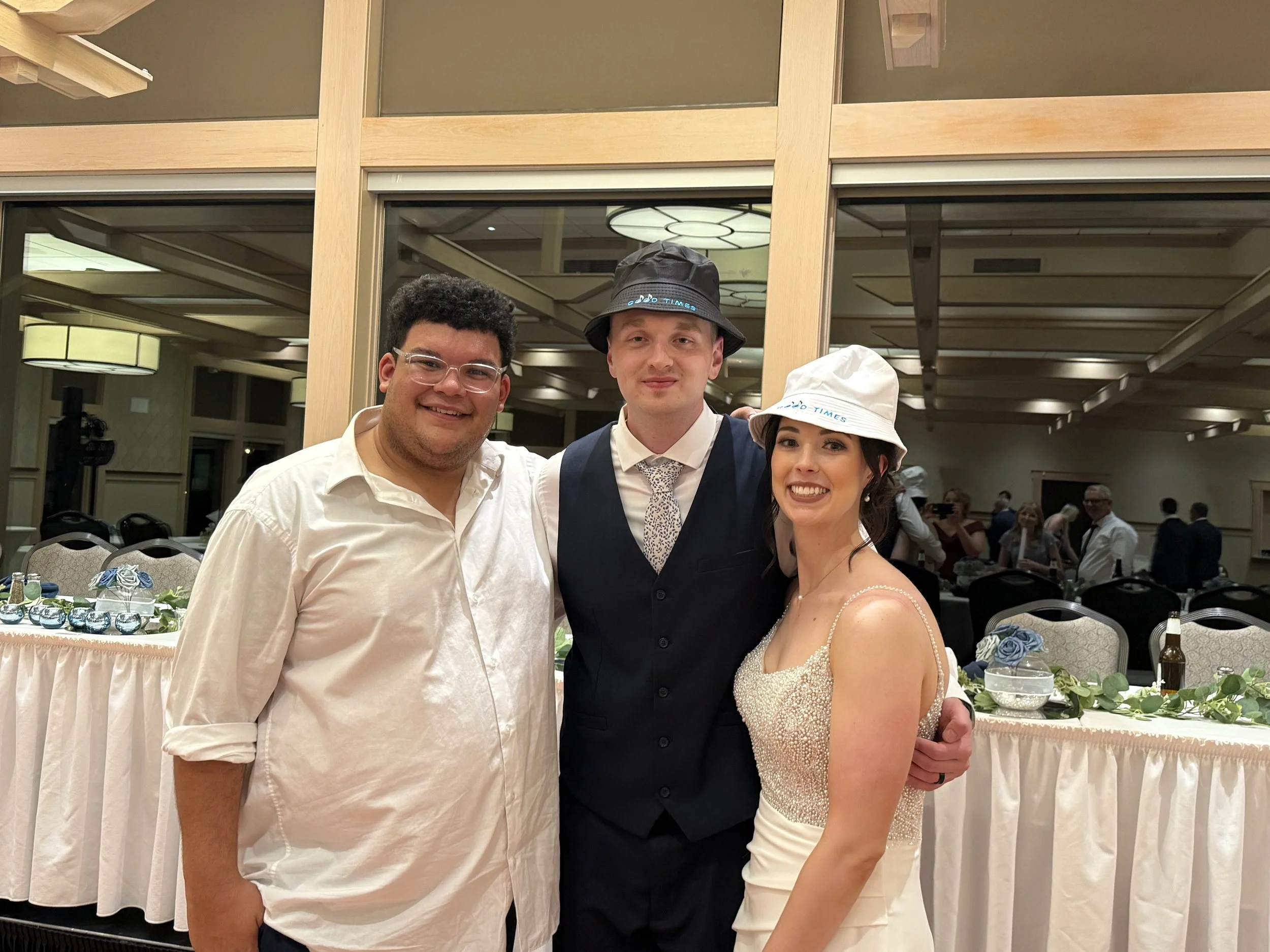 Three people dressed for a wedding or formal event standing together inside a decorated banquet hall, smiling at the camera.