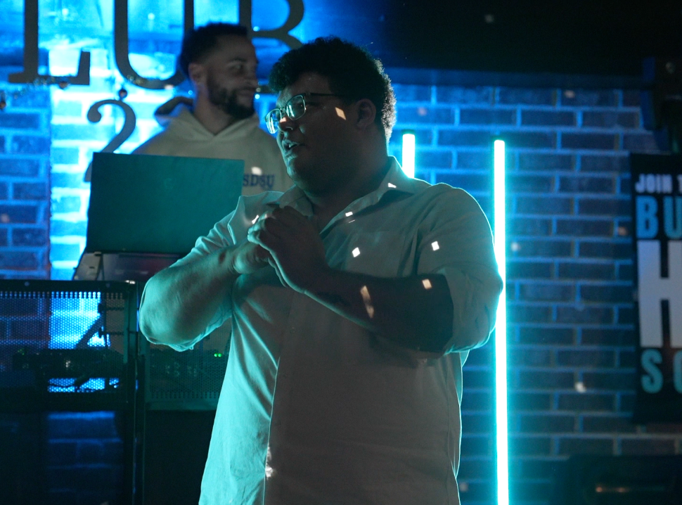 DJ Logan in the foreground with hands clasped on a microphone, wearing glasses and a light-colored shirt, in a dimly lit space with neon lights and brick wall background, while another person is in the background near DJ equipment.