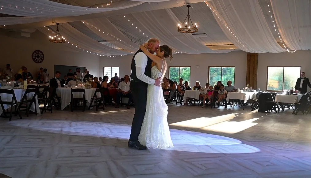 A bride and groom dance together at their wedding reception in a decorated hall with string lights and chandeliers, surrounded by seated guests.