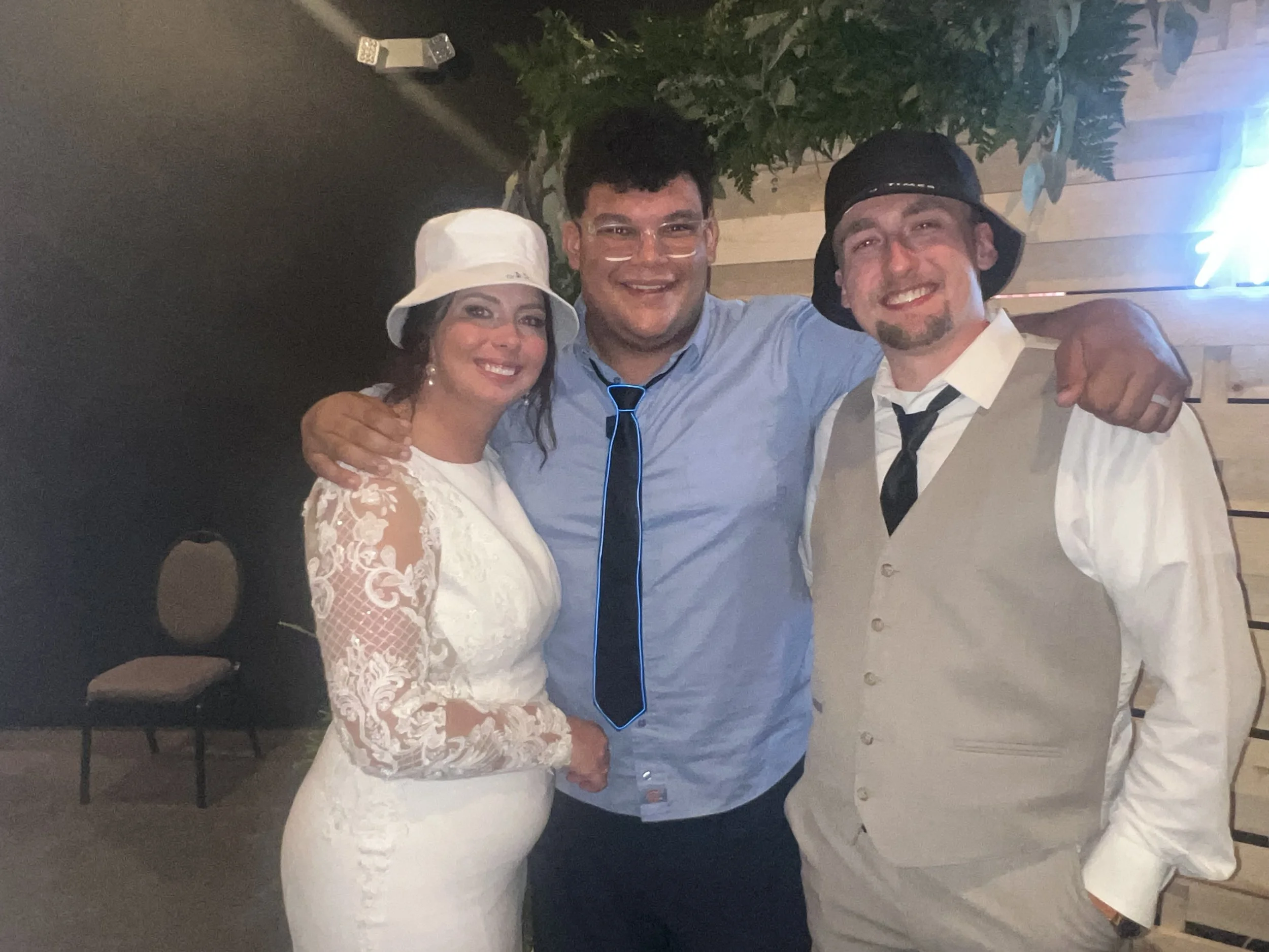 Three people smiling, standing close together with their arms around each other. A woman on the left is dressed in a white lace dress and a white hat. The man in the center is wearing a blue shirt with a black tie. The man on the right is dressed in 