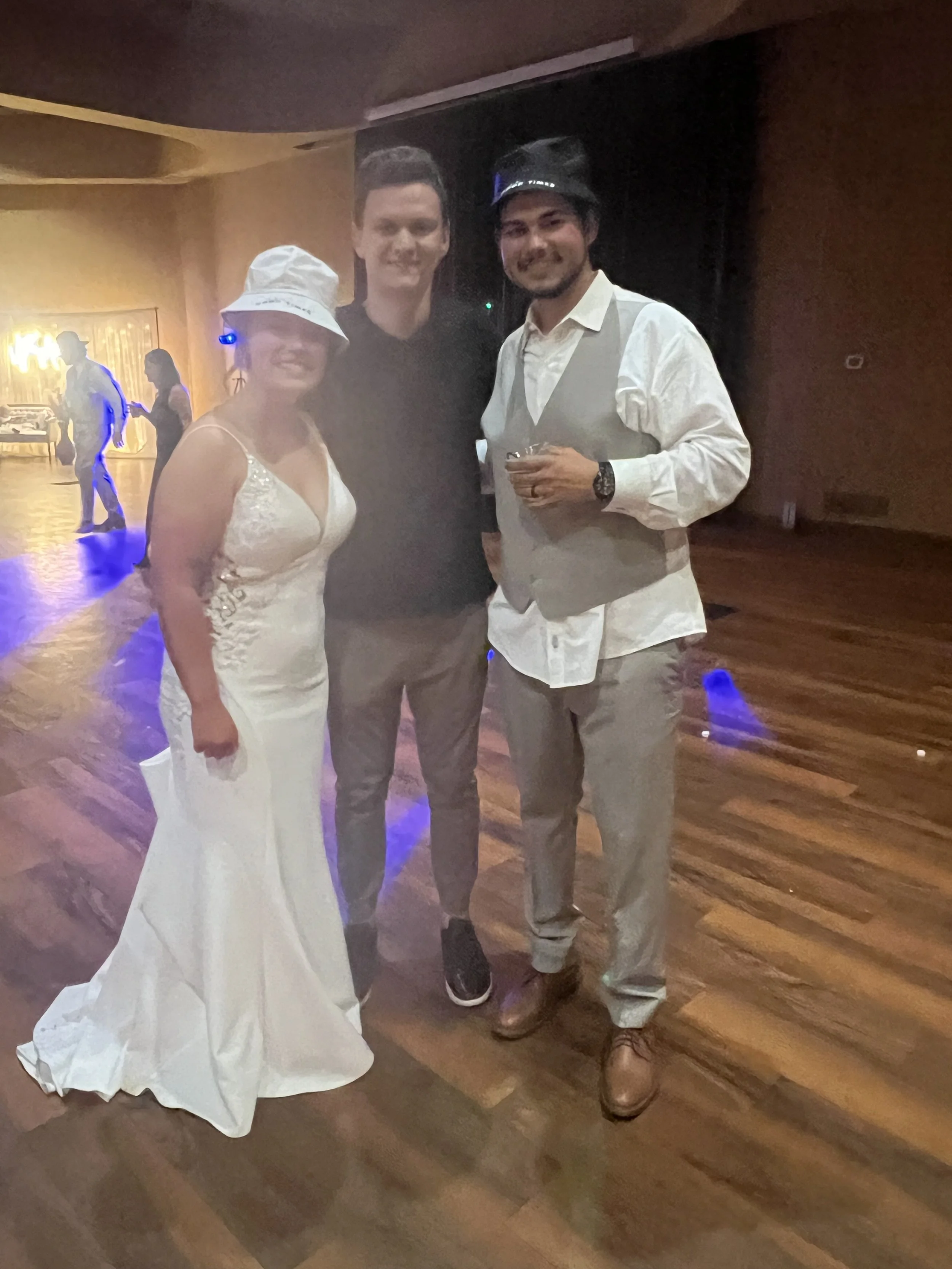 Three people at a party or celebration, dressed in semi-formal attire, on a wooden dance floor with colorful lighting.