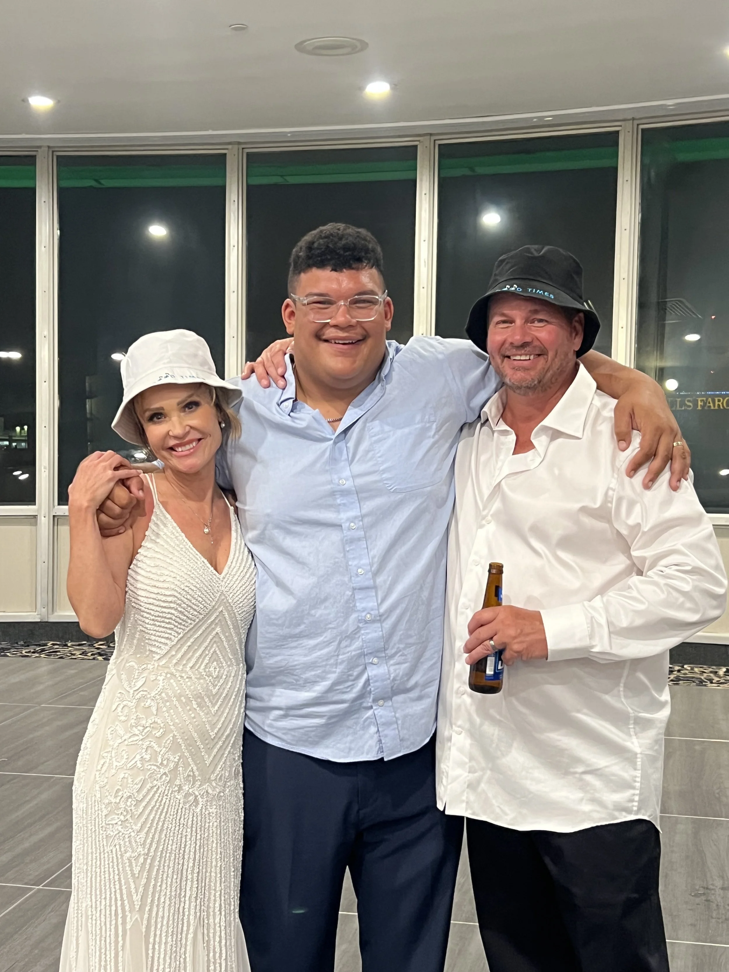 Three people smiling and hugging together indoors at night, with a woman on the left wearing a white dress and hat, a man in the middle wearing glasses and a light blue shirt, and another man on the right wearing a white shirt and black bucket hat ho
