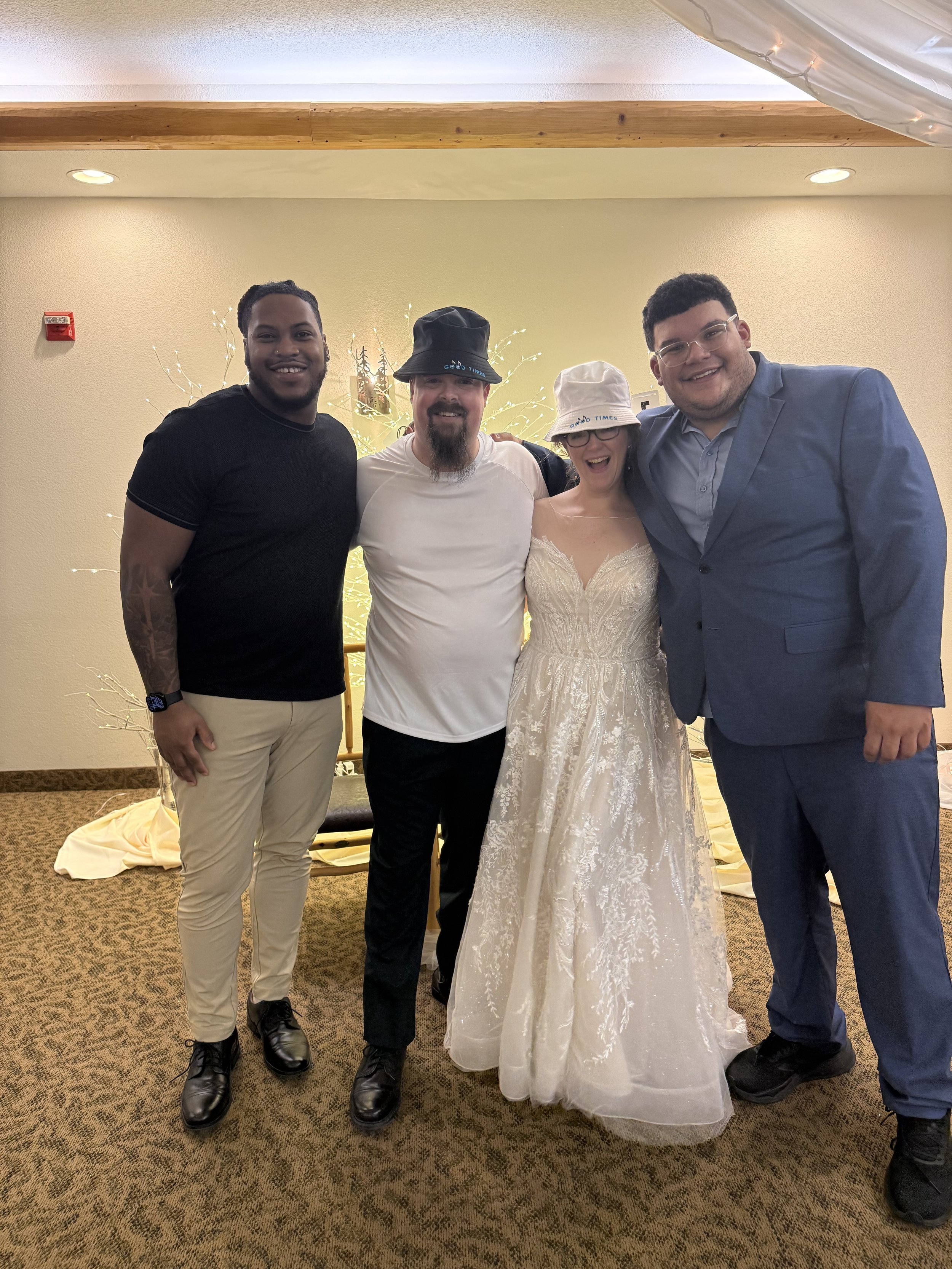 Group of five people standing together at a celebration, with a woman in a wedding dress at the center, all smiling and posing indoors.