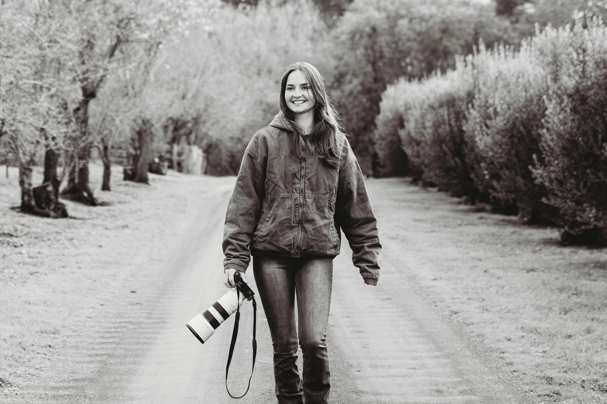 Black and white photo of a young woman smiling and walking outdoors on a dirt path, holding a camera with a large lens, surrounded by trees and bushes.