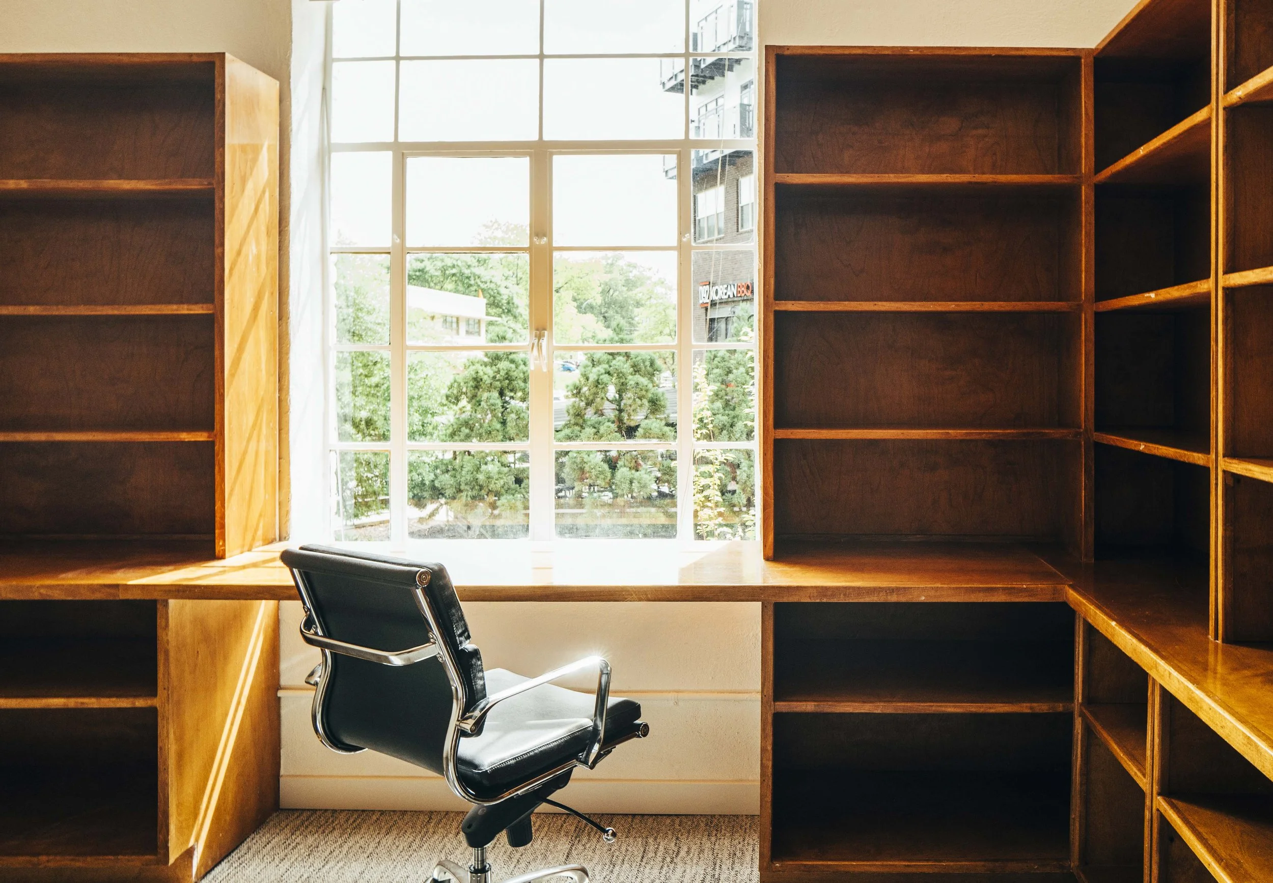 Empty office with built-in wooden bookshelves, a black office chair, and a large window with white framing and a view of trees and a building outside.