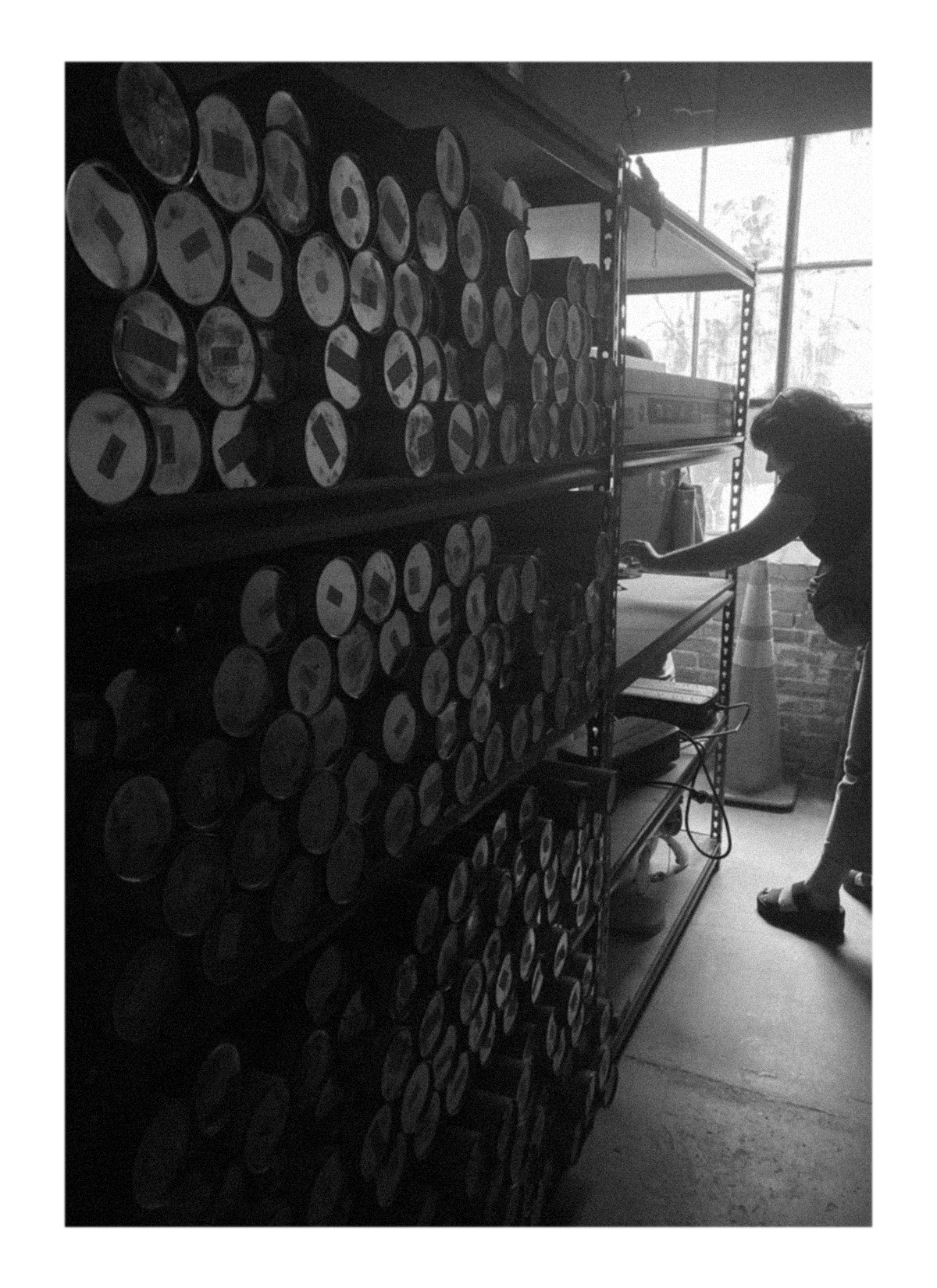 A person stands next to shelves filled with rolls of paper or fabric, using a tool, with a large window and a traffic cone visible in the background.