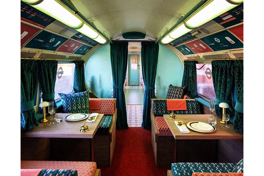 Inside a vintage train carriage with two dining tables set for two, decorated with colorful curtains, cushions, and tableware, under a curved ceiling with overhead storage.