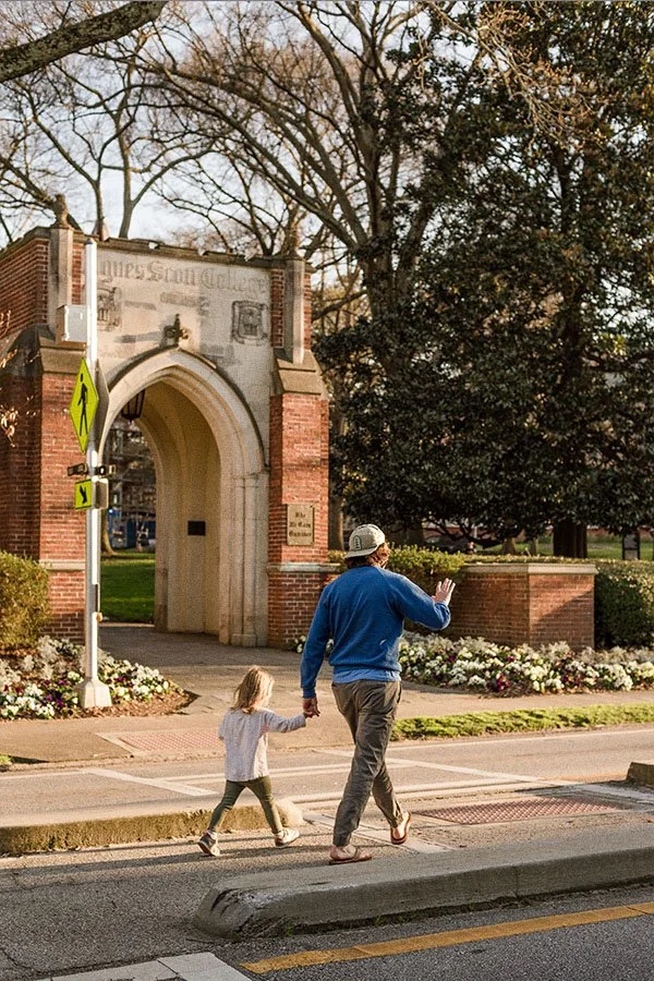 A man and young girl crossing a street holding hands in front of a brick archway entrance to a park or campus, with trees and flowers nearby.