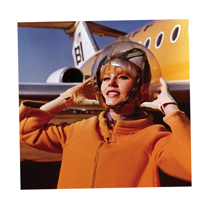 Woman in orange hoodie wearing a pilot's helmet standing in front of a yellow aircraft, adjusting her helmet as she prepares for flight.