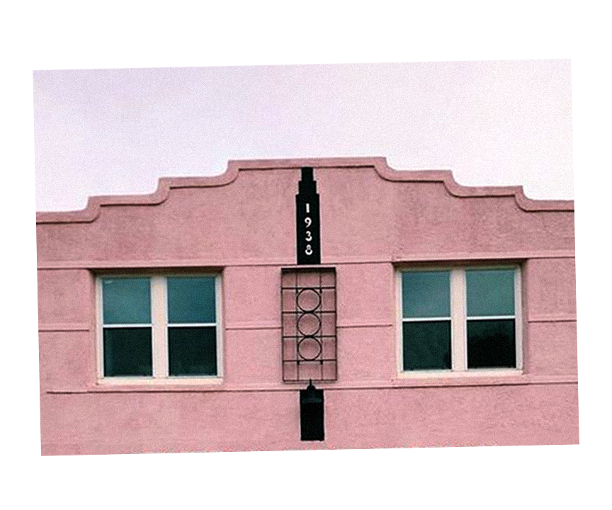 Pink building facade with two symmetrical windows, decorative vertical black element with the year 1938, and a geometric design resembling a clock or window frame.