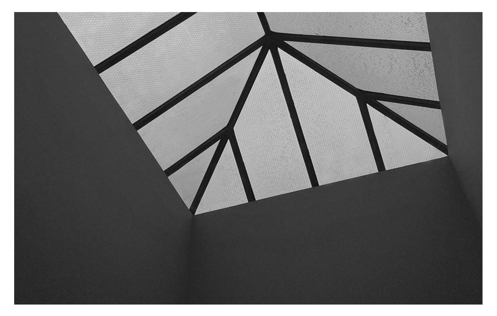 Black and white photograph of a geometric skylight ceiling with metal framing and glass panes, viewed from below.