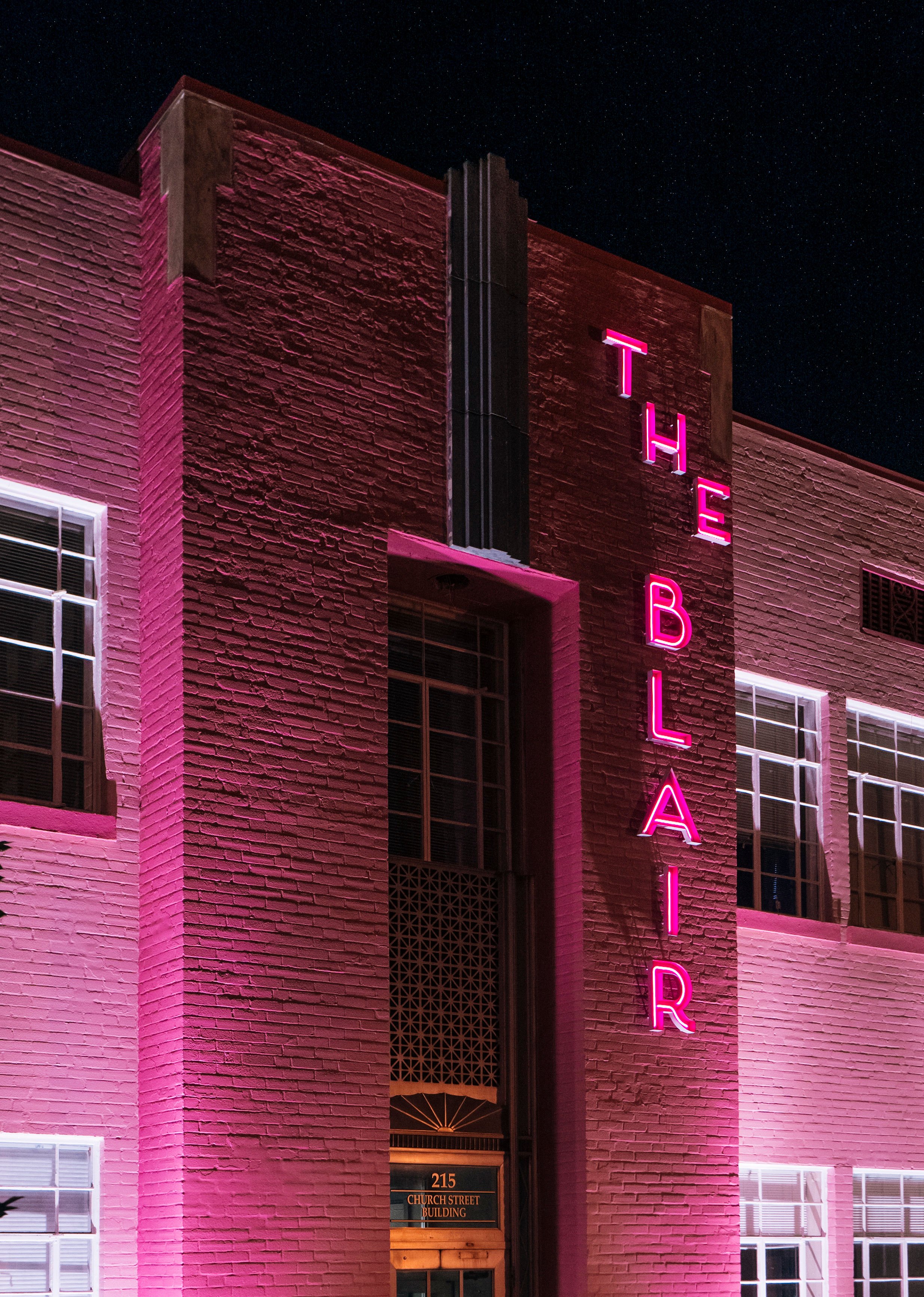 Night view of a brick building with a neon sign that reads 'THE BALLET' in pink letters. The building has multiple windows lit from within and a small sign at the entrance reading '215 CHURCH STREET BUILDING'.