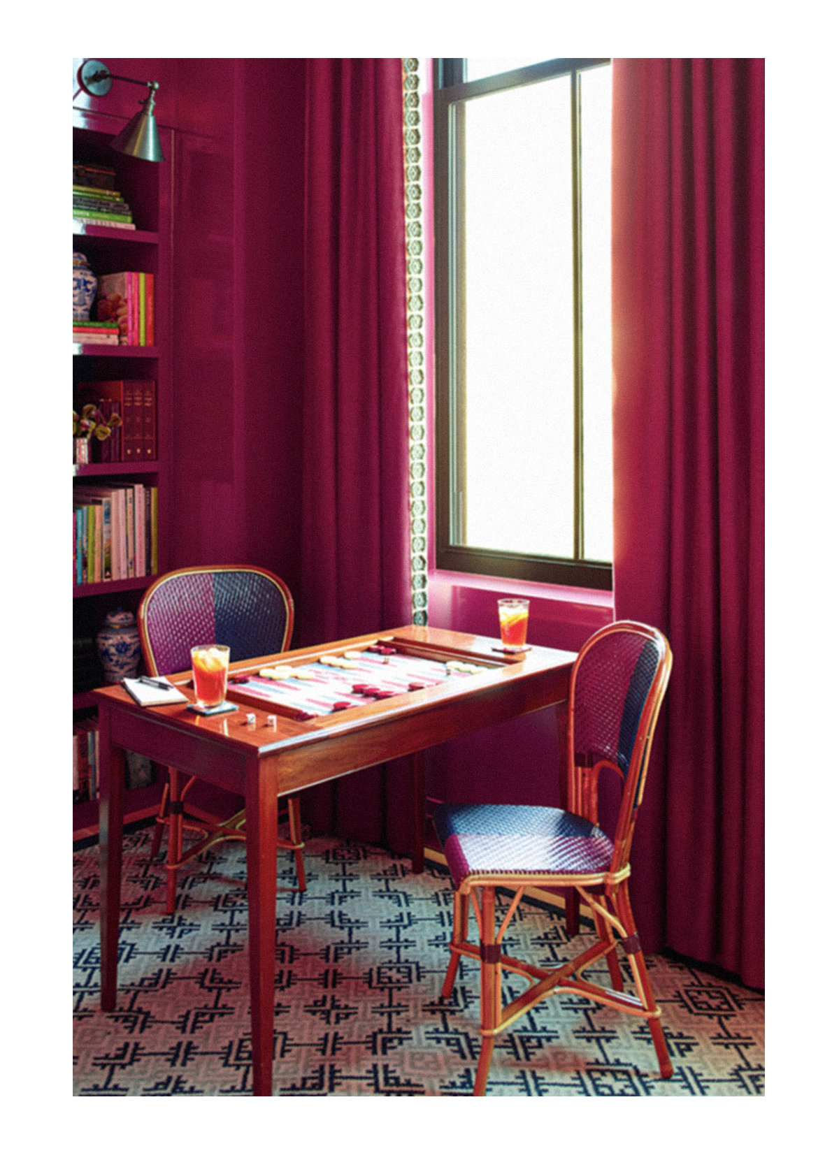 A cozy room with dark pink walls, a window with sheer curtains, a wooden table with tabletop games and two drinks, and two chairs with pink and dark blue upholstery. There is a bookshelf filled with books and decorative items on the left side.