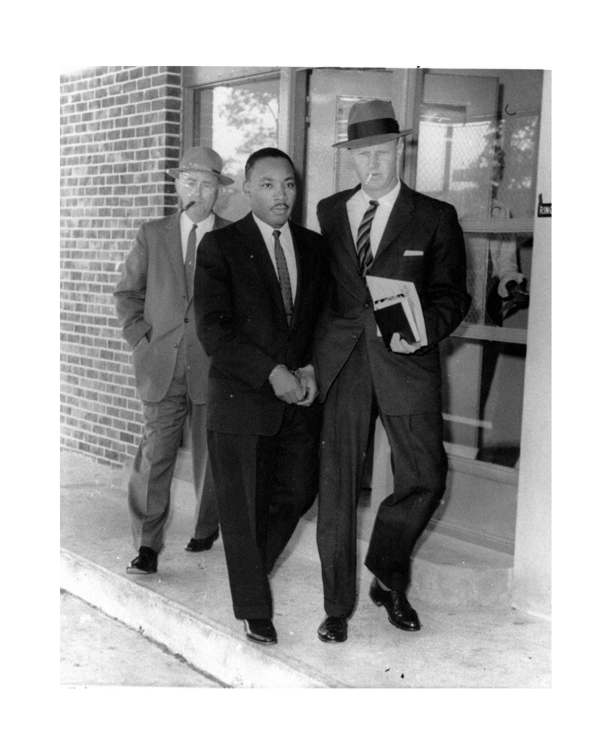Black and white photo of two men in suits shaking hands outside a building, with another man in a suit and hat standing behind them.
